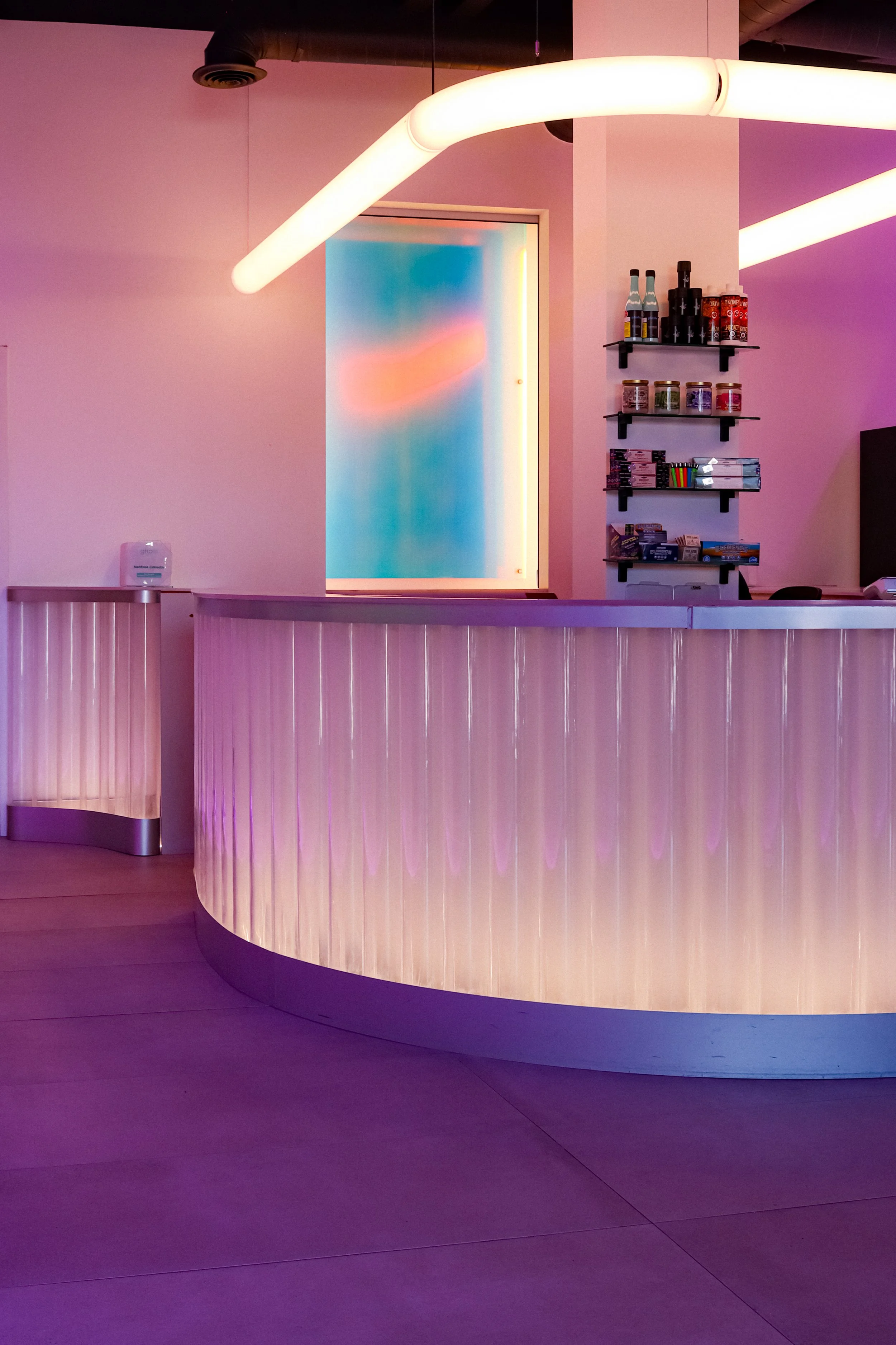 A modern, curved reception desk made of light-colored wood, with a vertical slat design, situated in a dimly lit room with purple and pink neon lighting. Behind the desk are three black shelves holding various bottles, jars, and boxes. There is a doo
