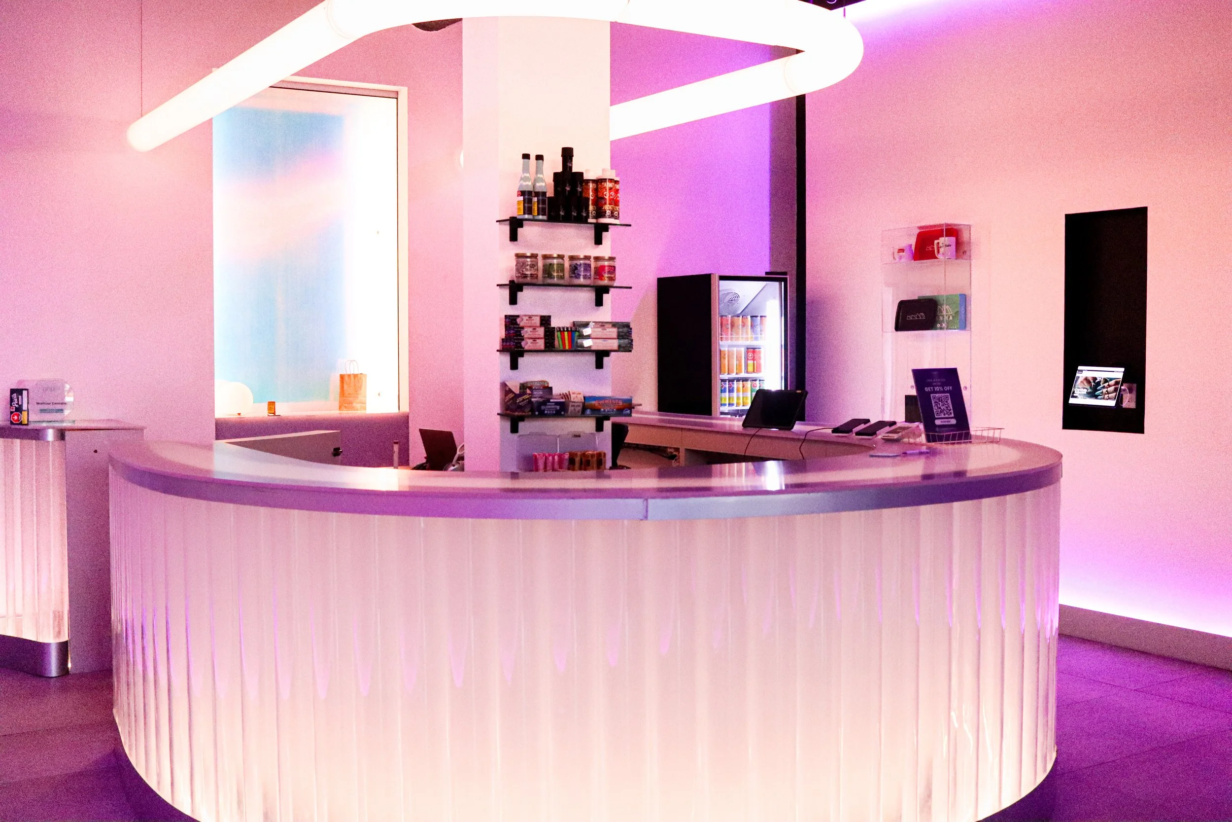 A modern food or drink counter with a curved pinkish-purple base, a white countertop, and a large overhanging light fixture. Shelves with bottles, jars, and books are mounted on a pink wall behind. A small refrigerator with beverages inside and a tou