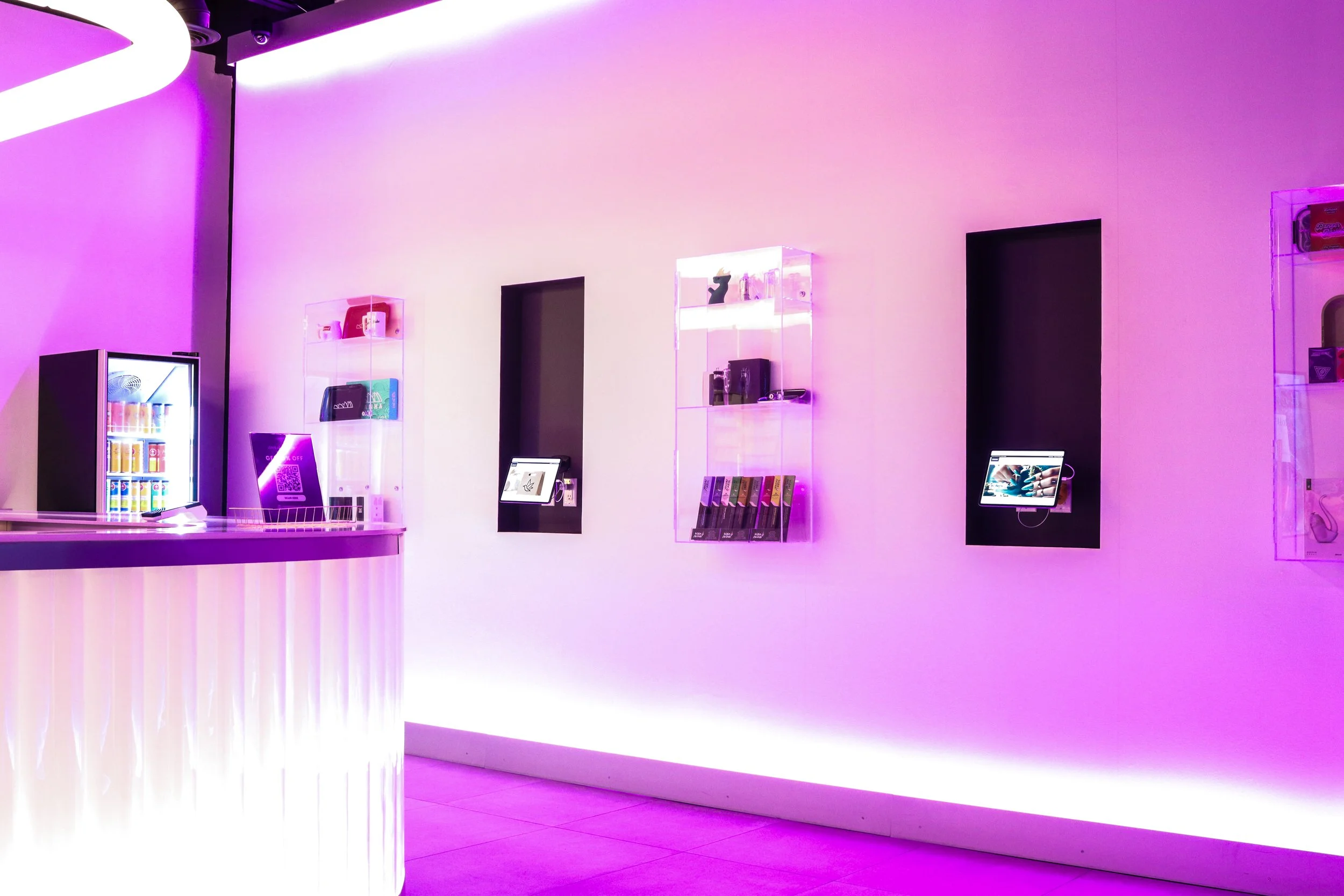 Interior of a store with pink and purple lighting, display cases with products, and digital screens