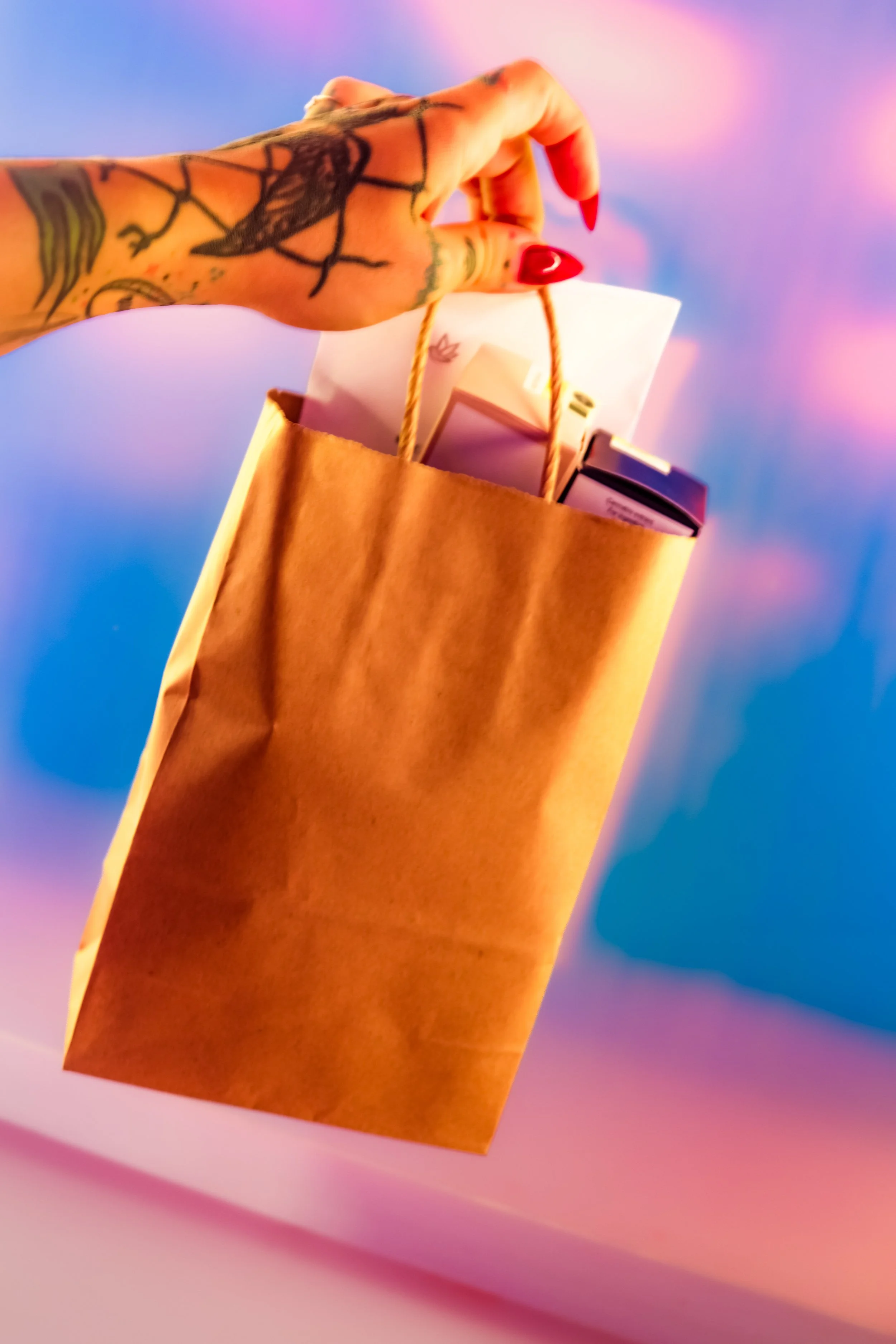 A hand with a tattoo holding a brown paper shopping bag with rope handles, against a pink and purple blurred background.