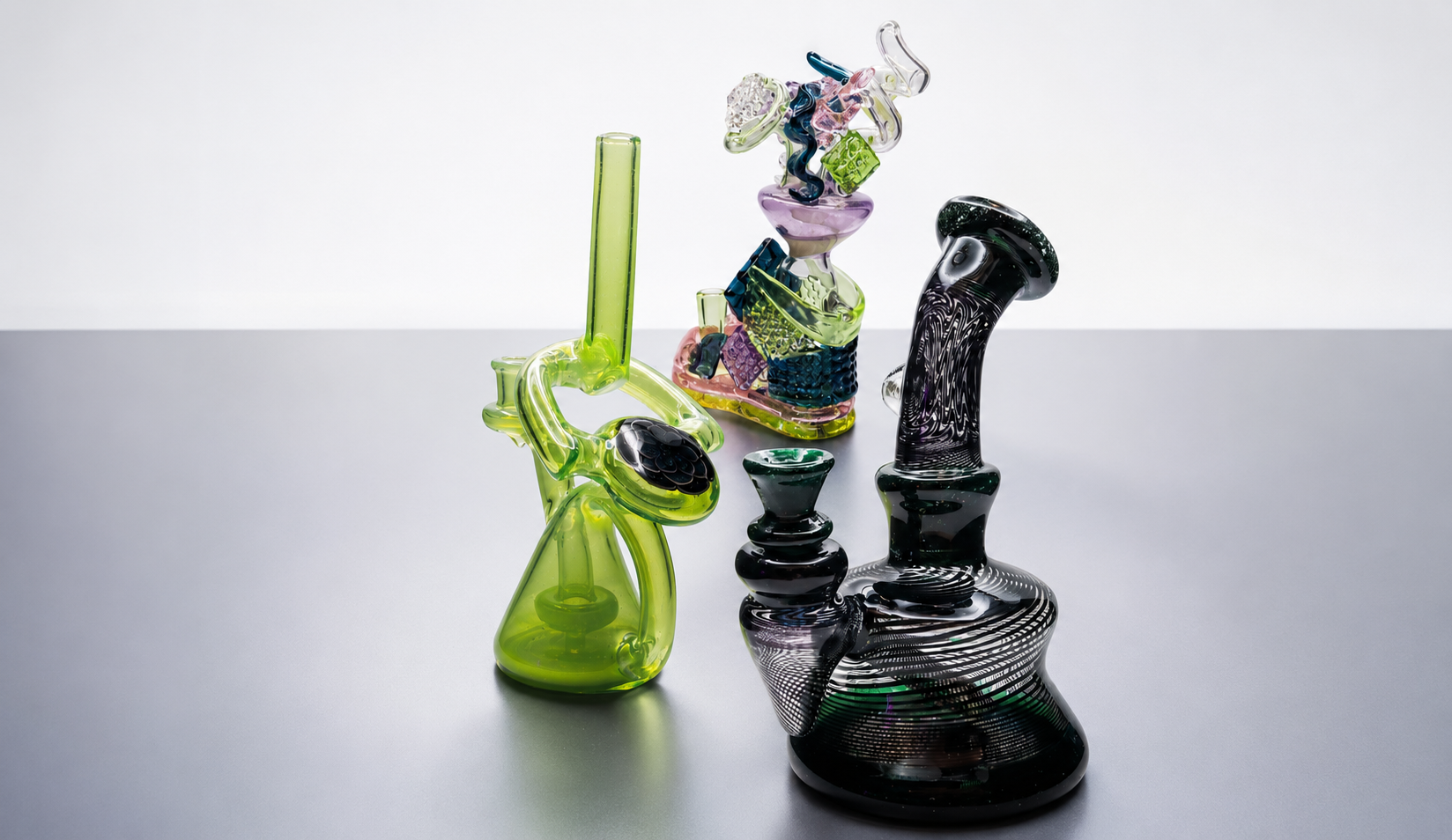 Two colorful glass smoking pipes with intricate designs and a multicolored glass sculpture in the background on a plain grey surface.