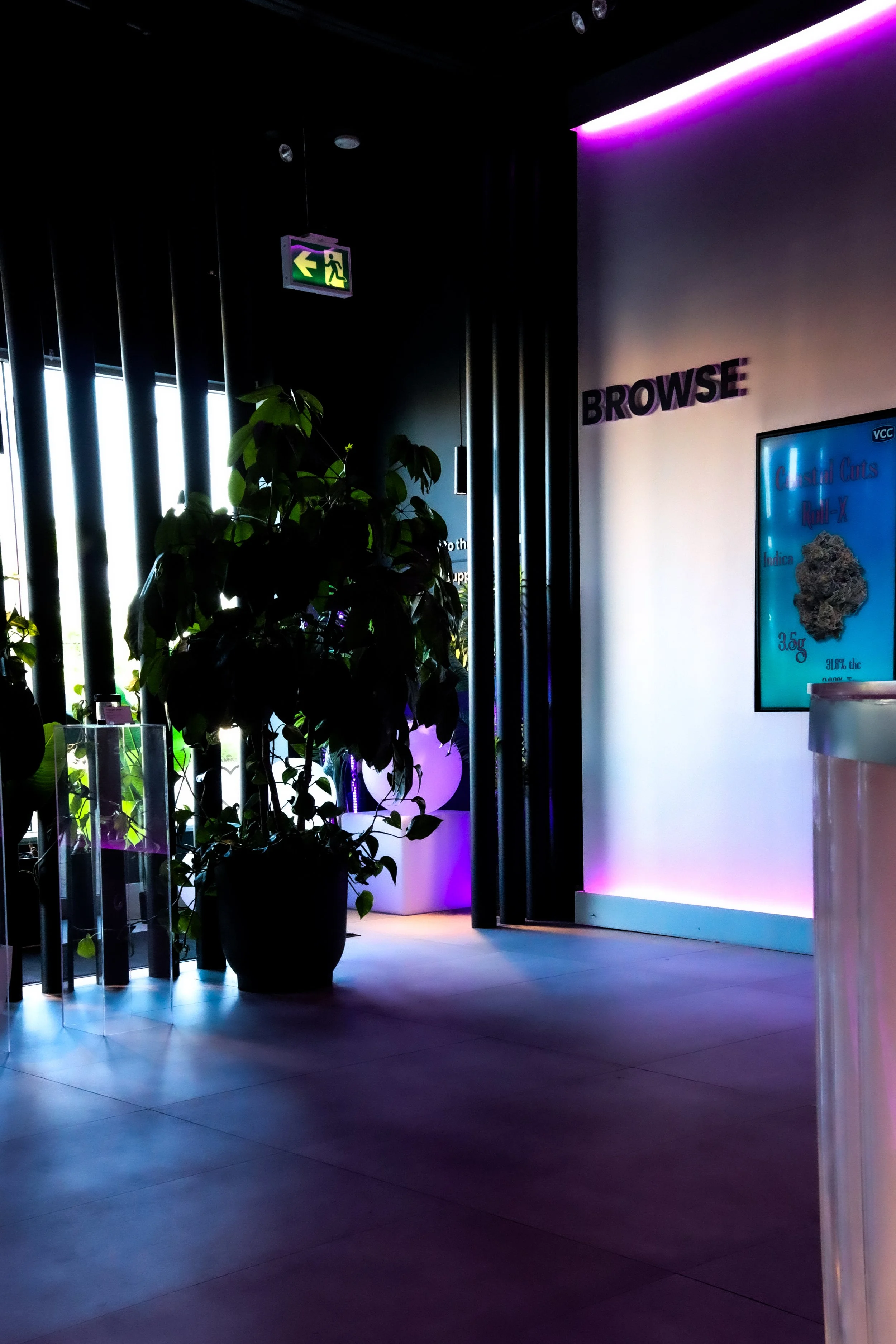 Interior of a modern establishment with black vertical slats, a potted plant, and purple accent lighting. An illuminated emergency exit sign with a left arrow and running figure is visible on the ceiling. Part of a digital menu or advertisement is vi