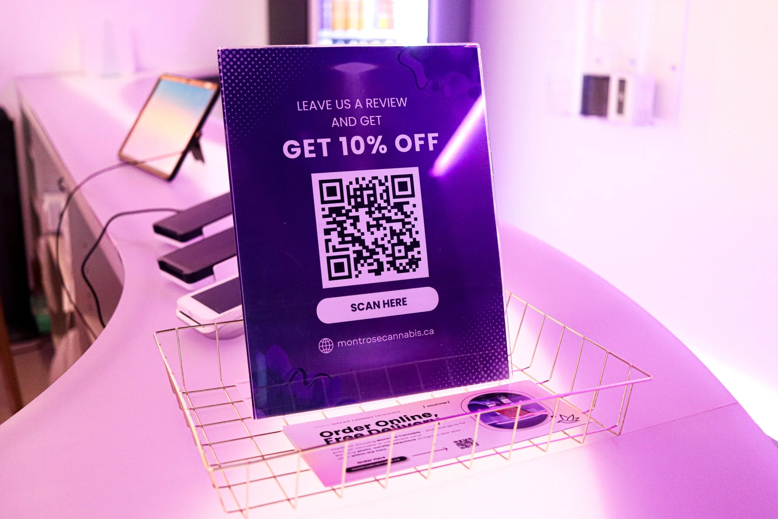 A purple promotional sign on a pink table offering a 10% discount for leaving a review, with a QR code below the text. The sign is placed in a gold wire basket, with a woman playing a keyboard and another person holding a tablet visible in the backgr