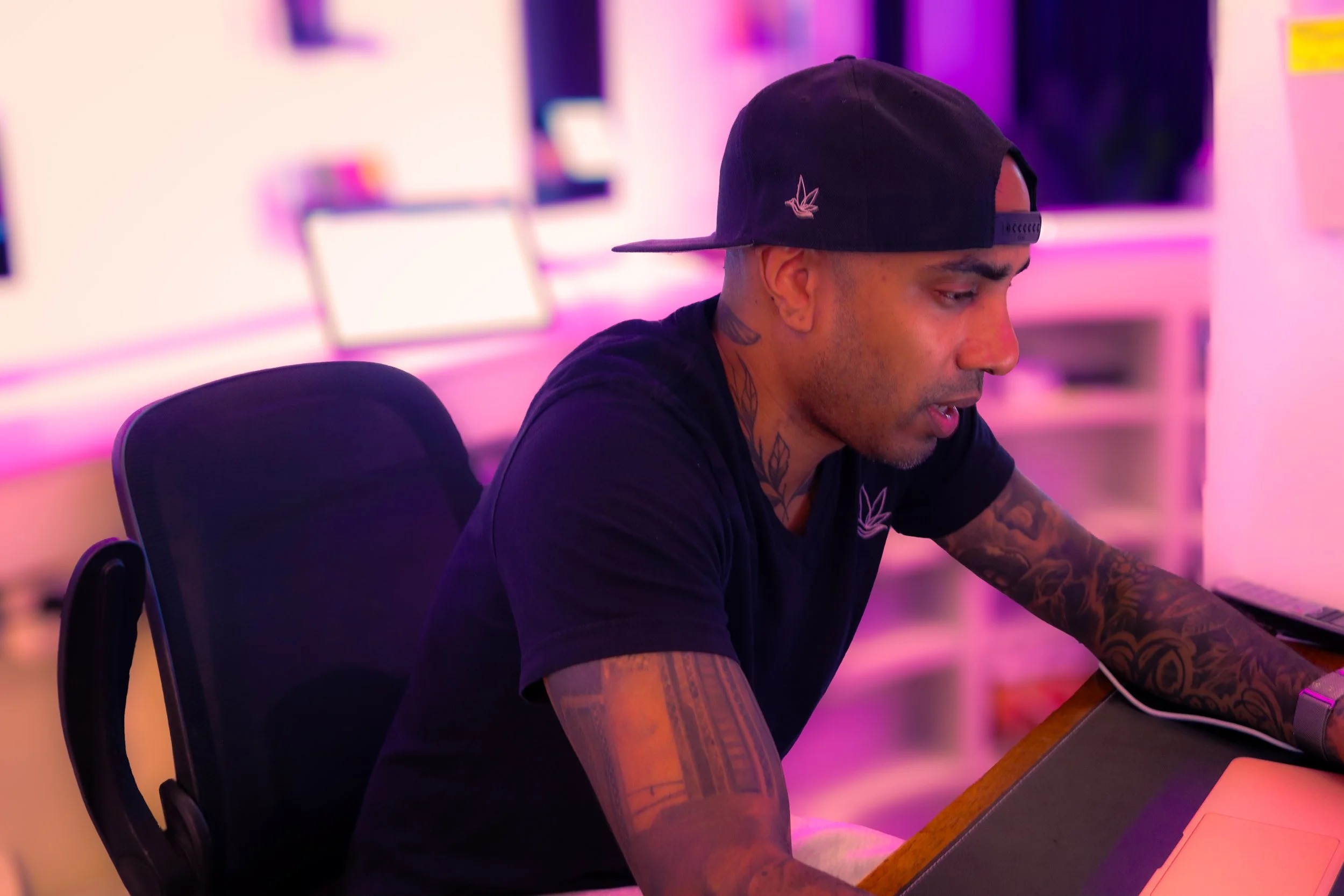 A young man with tattoos and a black baseball cap sitting at a desk in a room with pink and purple lighting, working on a computer.