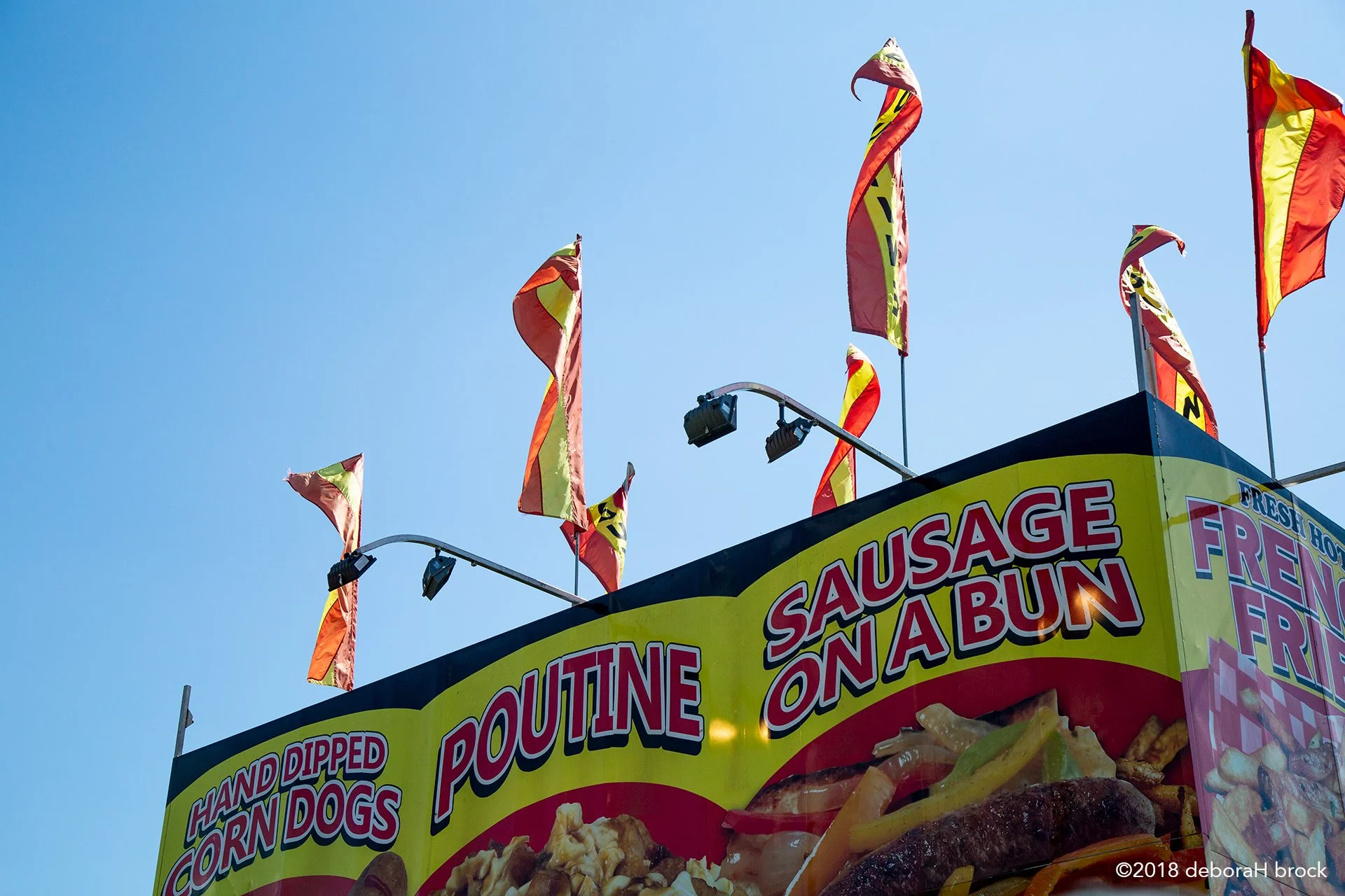 A food stand with yellow signage advertising sausage on a bun, corn dogs, and fried foods. Several red and yellow flags are flying against a clear blue sky.