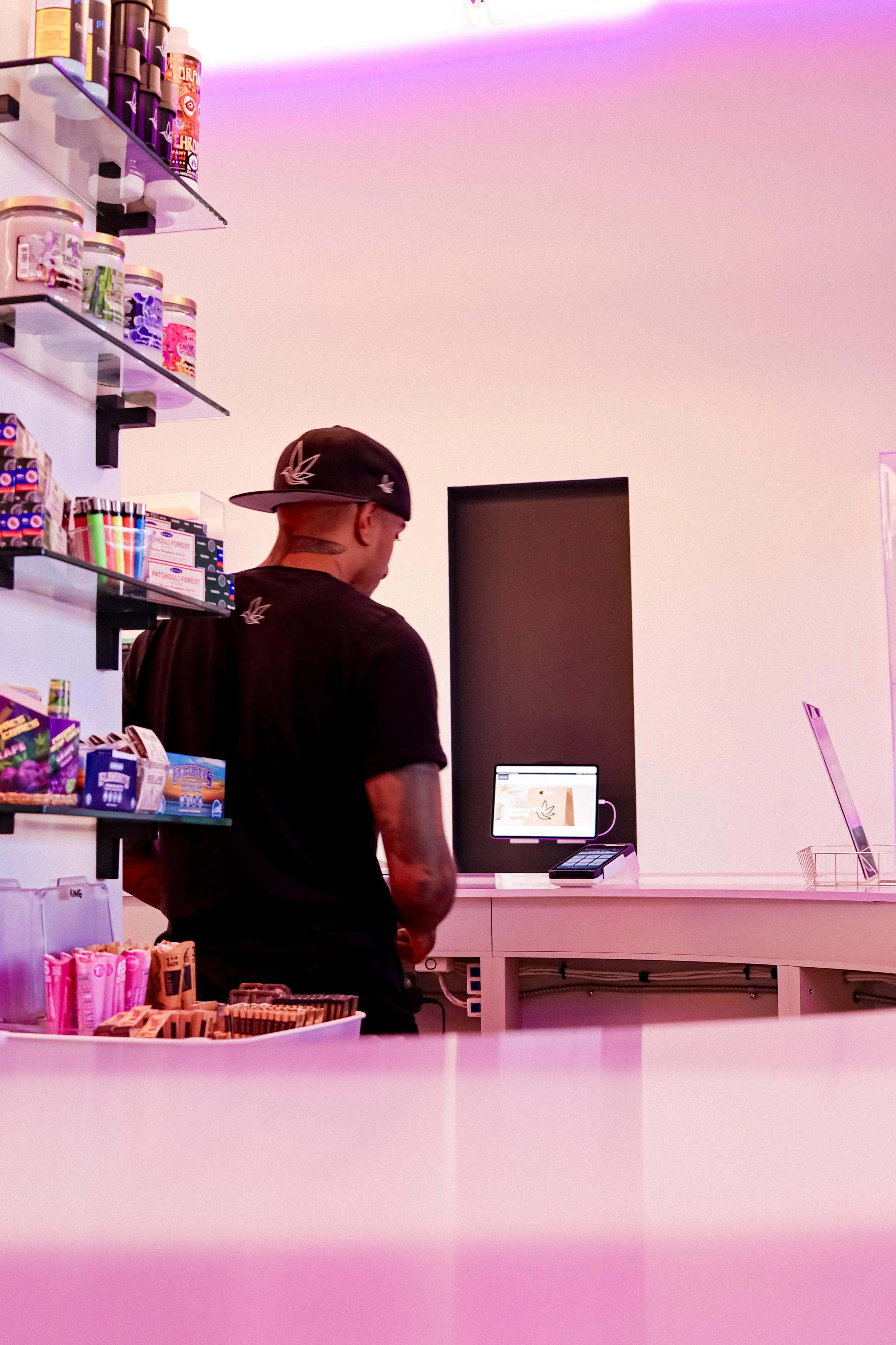 A person wearing a black baseball cap and black t-shirt with a leaf logo, standing at a checkout counter in a store. The store has shelves with snacks and colorful items, and there is a pink and purple lighting ambiance.
