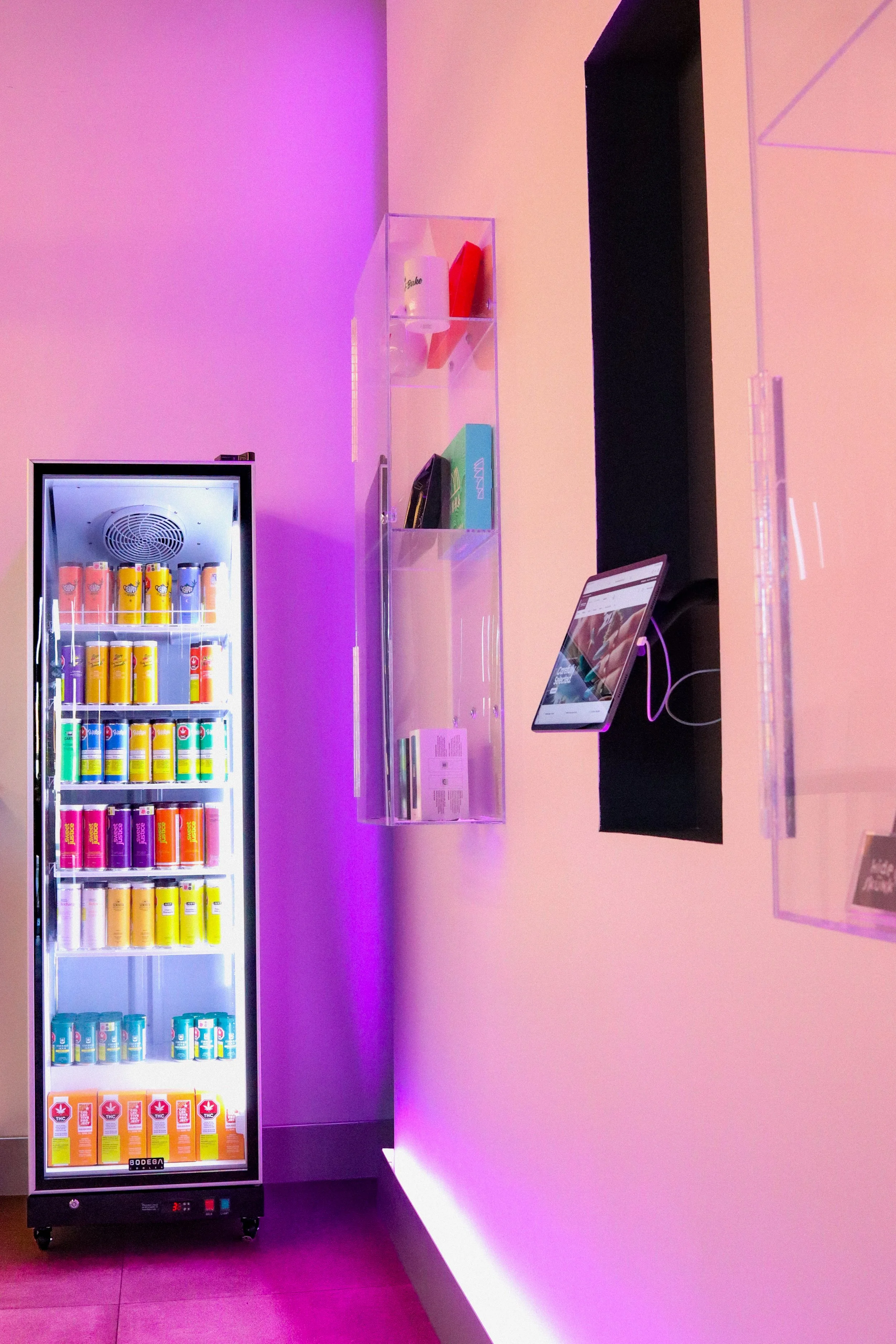 A refrigerated display case filled with colorful cans of beverages, situated next to a purple wall with a pink gradient. To the right, there is a black counter with a computer monitor attached to a pink wall, illuminated by purple and pink lighting.