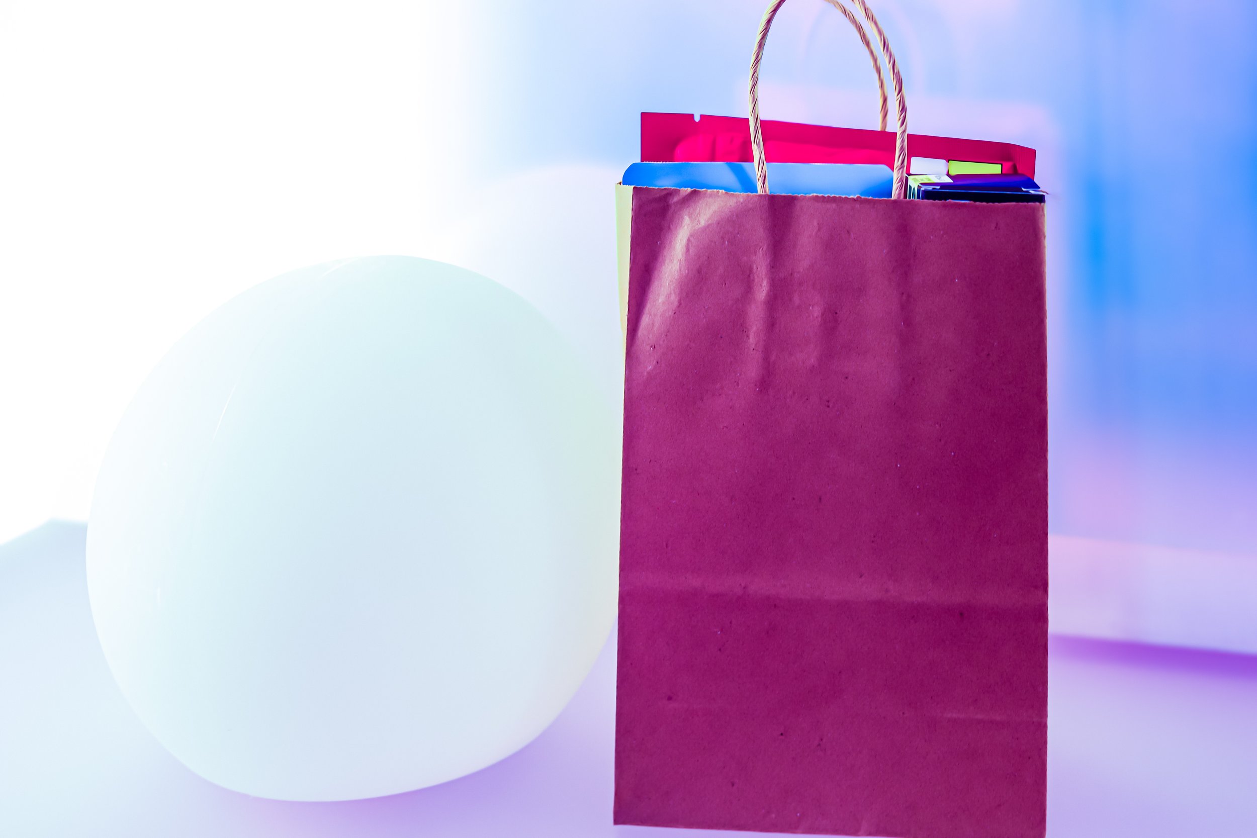 A store shopping bag in maroon color with rope handles containing colorful boxes or packages, against a soft gradient background.