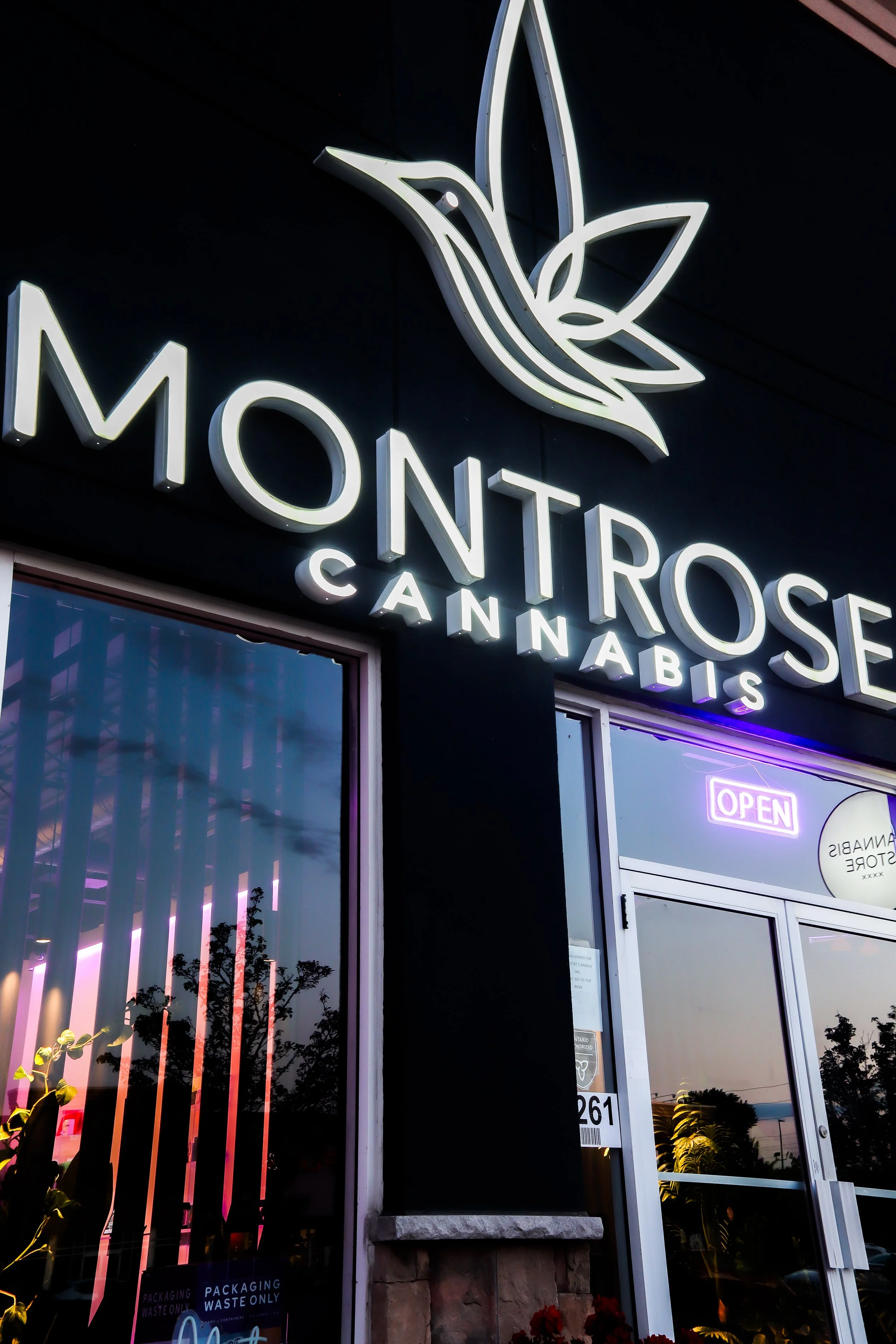 Exterior view of a cannabist store called 'Montrose Cannabis,' with illuminated signage, a large stylized leaf logo, and an 'Open' sign near the entrance, reflecting sunset and greenery outside.