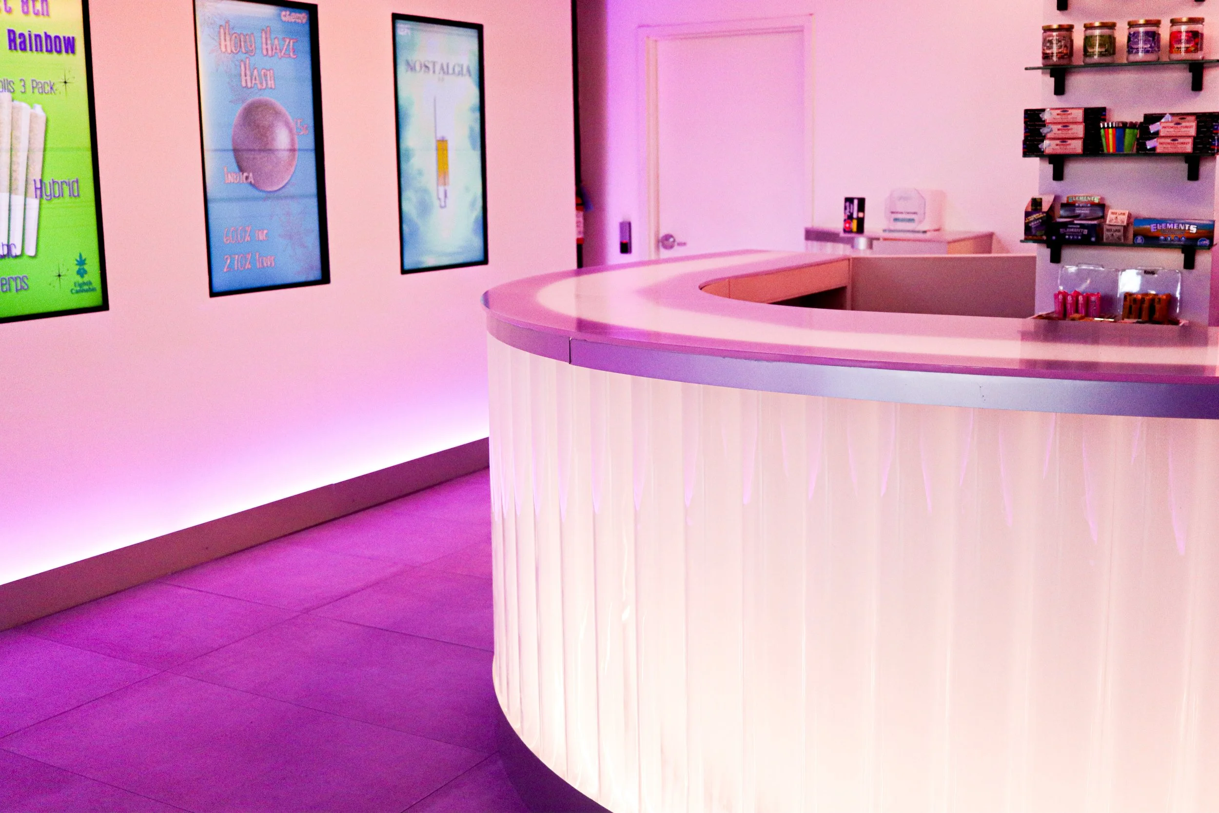 A pink reception counter with vertical lighting details, set in a room with pink walls and a purple-lit ceiling. On the walls, there are colorful posters, and shelves with various products are visible in the background.