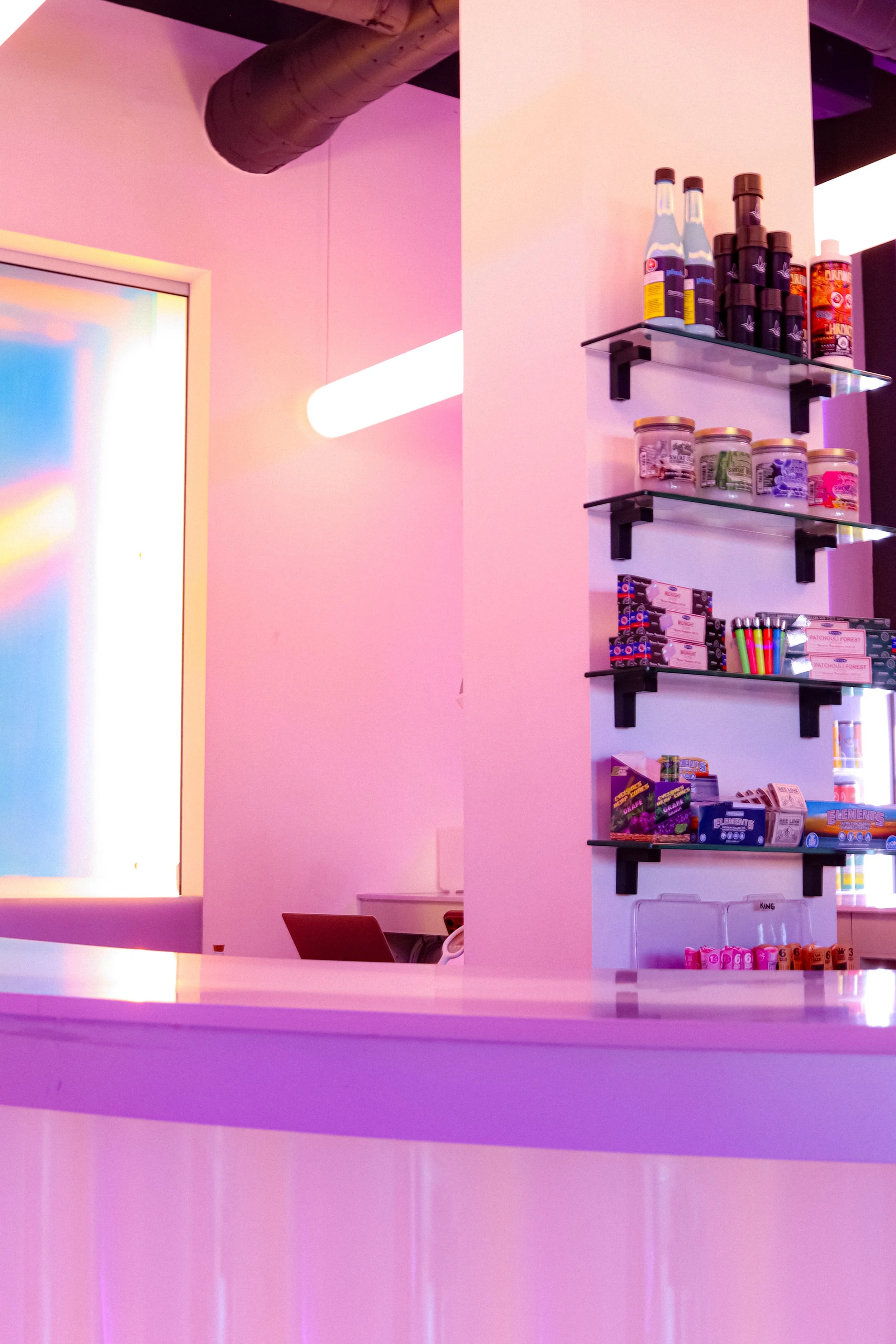 Pink wall with shelves holding various bottles and containers, colorful items, and a neon light.
