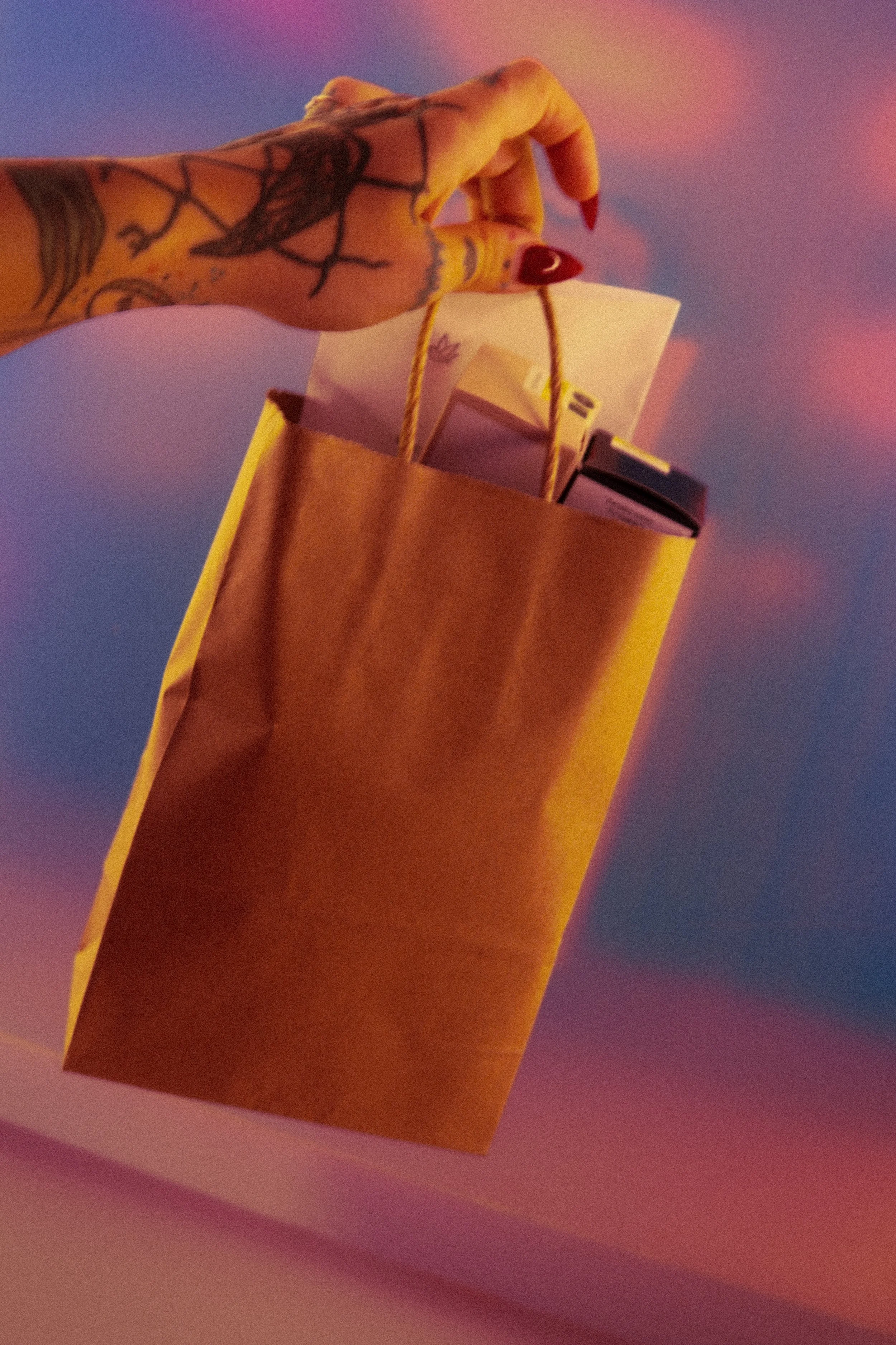 Close-up of a hand with tattoos holding a small paper shopping bag with yellow handles. The background is a pastel-colored gradient.