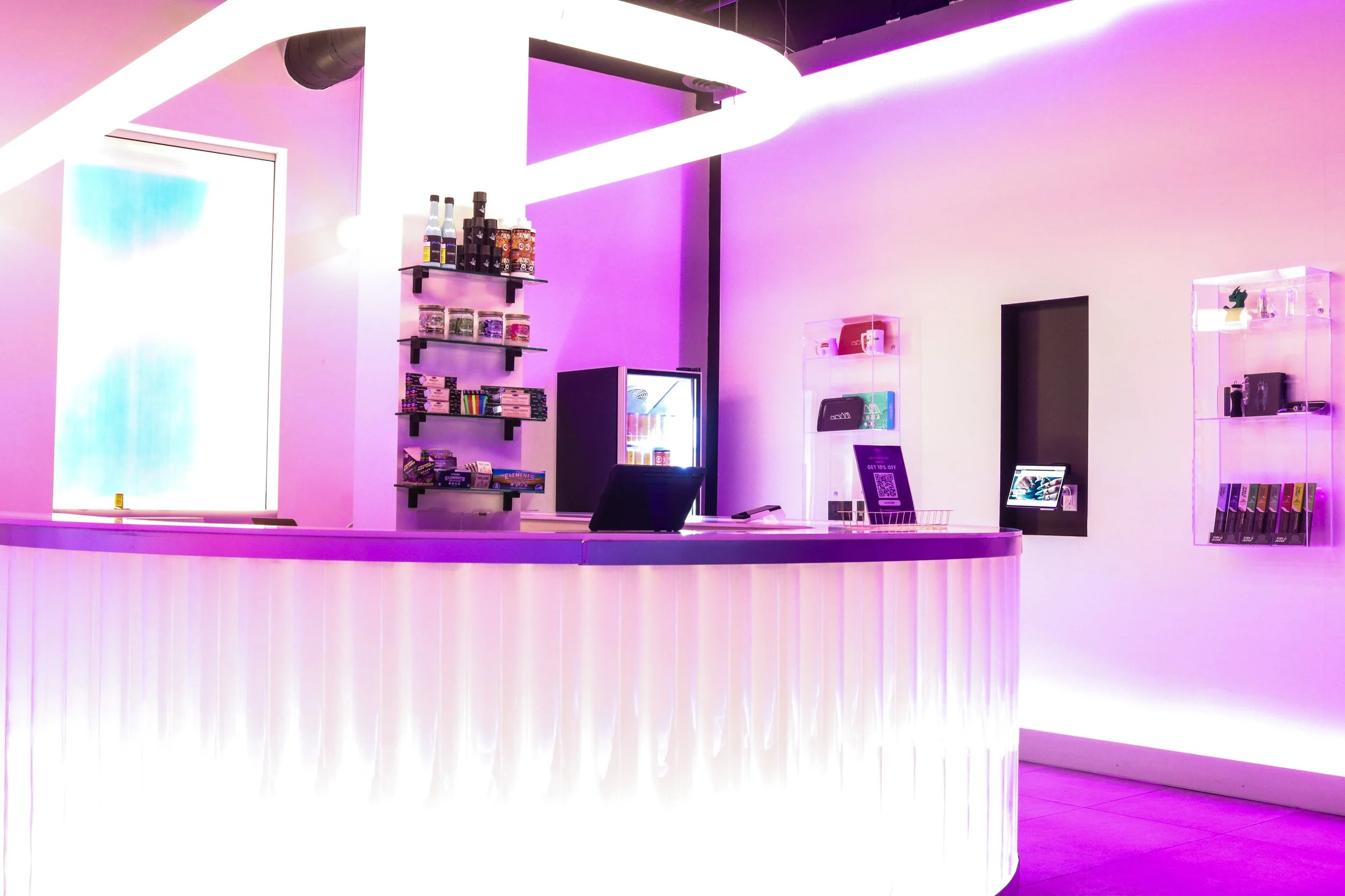 A brightly lit reception area with a curved white counter, pink and purple lighting, shelves with various products, and a small refrigerator behind the counter.
