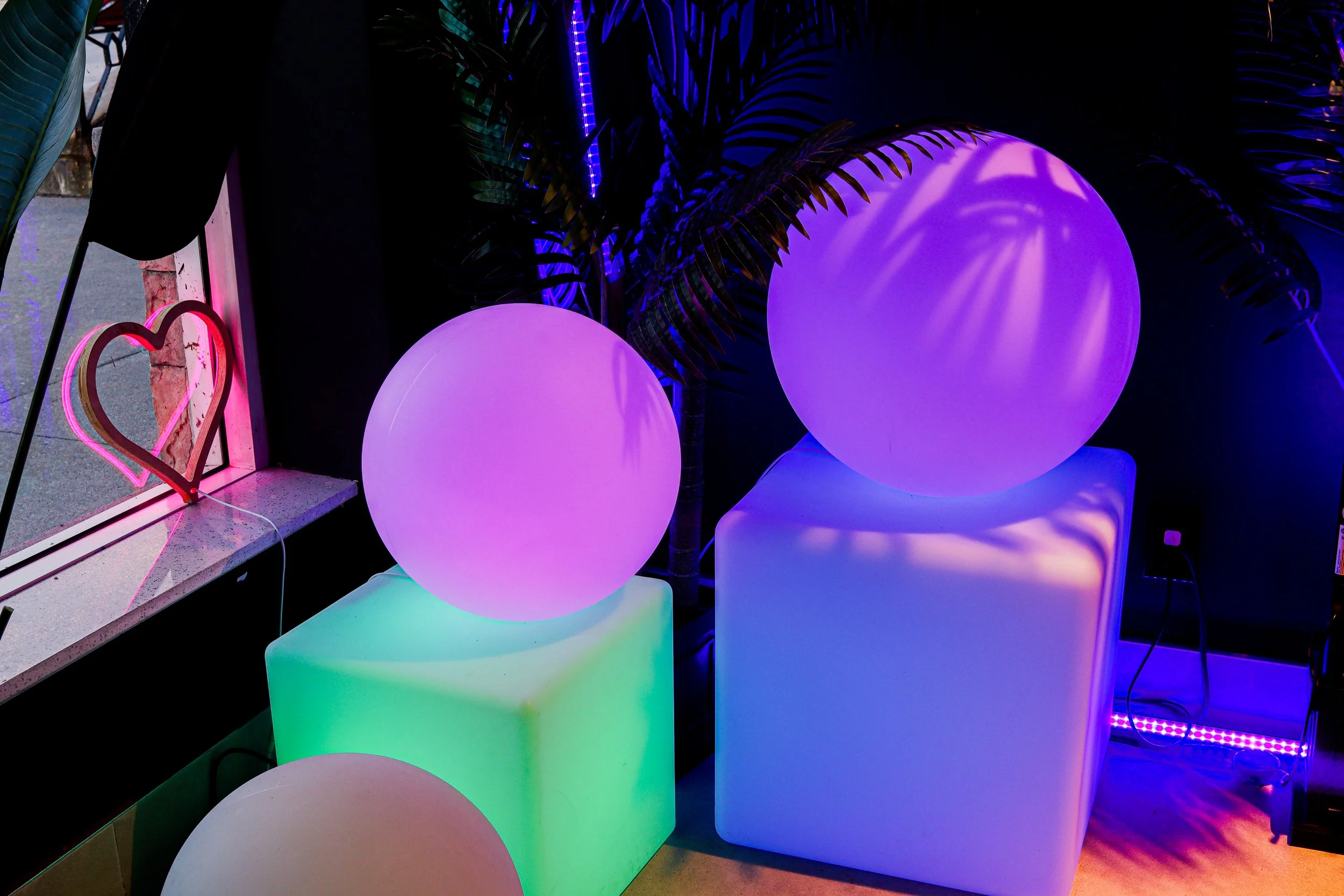 The image shows illuminated spherical and cube-shaped objects glowing in various colors including purple, green, and pink. There are decorative palm leaves and a neon heart sign near a window with a pink border, creating a vibrant, colorful atmospher
