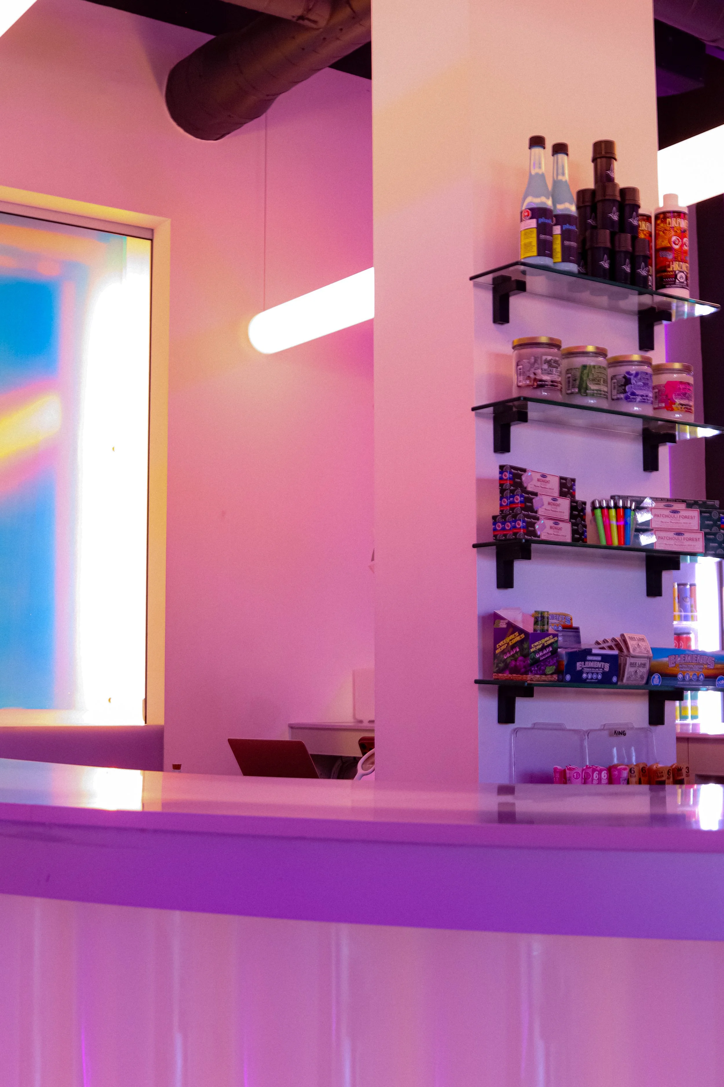 A purple-lit convenience store corner showing shelves with bottled drinks, jars of candies, and packaged snacks, with a window and neon light in the background.