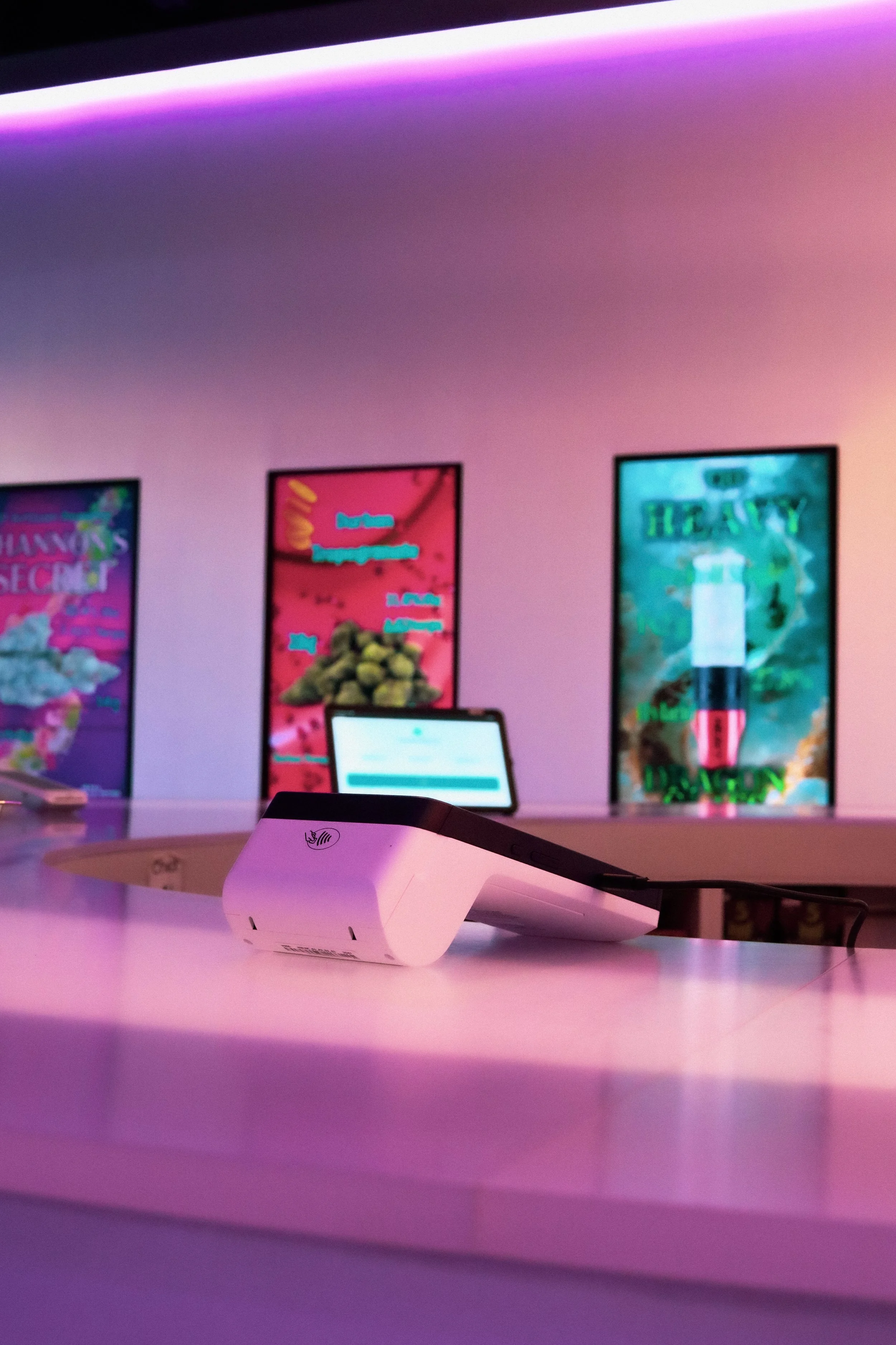 A white electronic device with a sticker and a black cable on a pinkish table. In the background, there are three vertical digital screens with colorful graphics. The ceiling has purple and pink lighting.