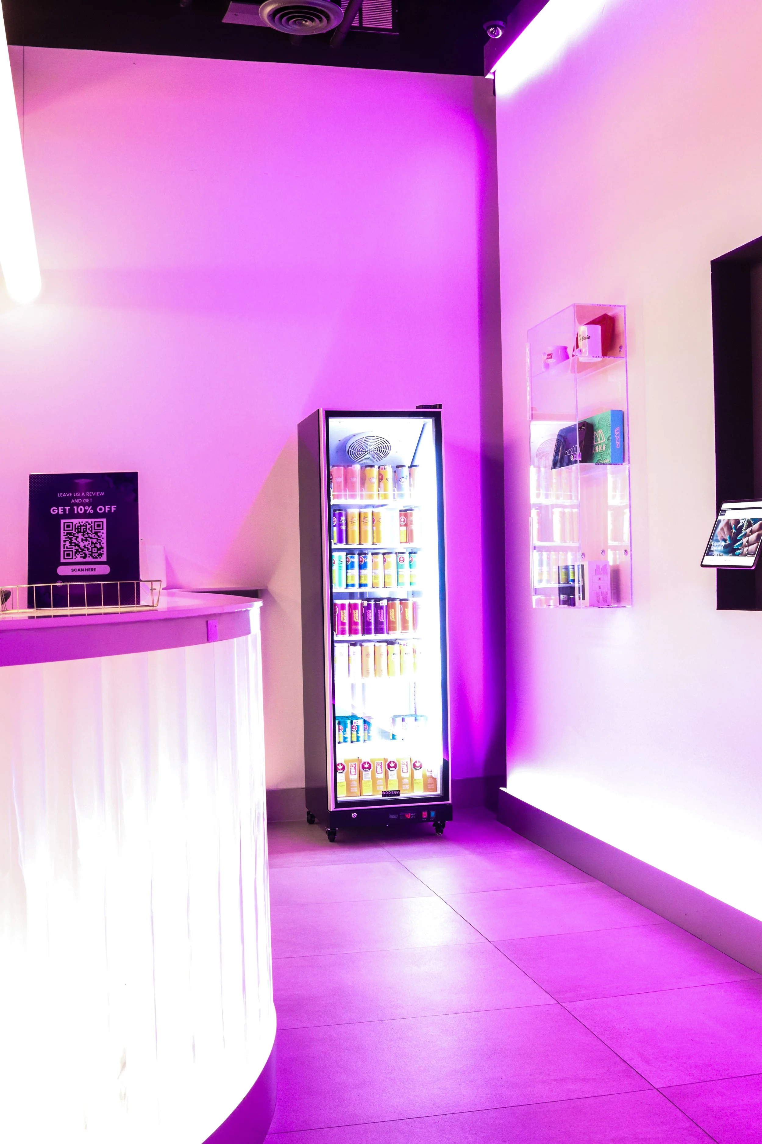 Interior of a modern, dimly lit space with purple LED lighting. There is a white curved counter on the left, a beverage refrigerator in the center, and a clear display shelf on the right with products. A QR code sign is on the counter.