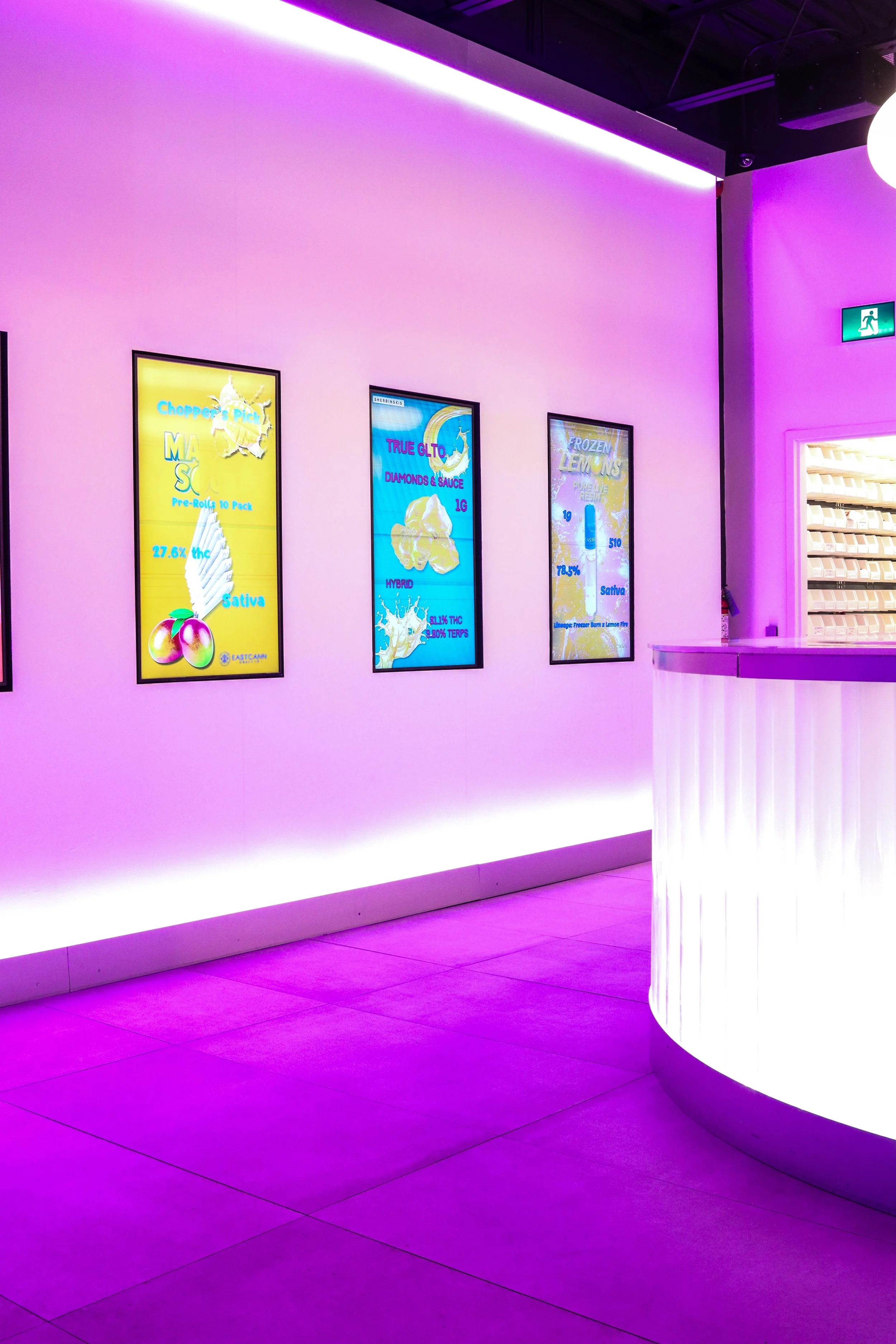 Interior of a store or lounge with a pink and purple neon-lit wall, digital menu boards displaying product advertisements, and a white display counter with vertical lighting. Exit signs are visible on the walls.