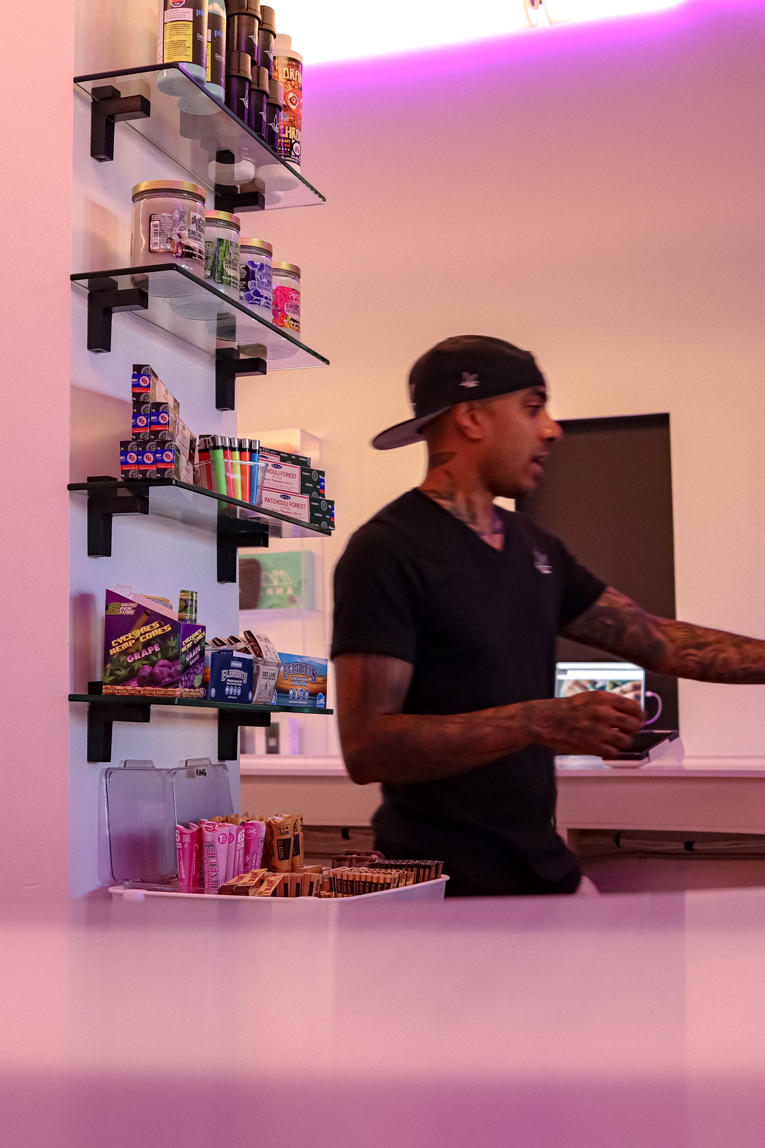 A man wearing a black baseball cap and black t-shirt is standing behind a counter in a store, holding a small white object. Behind him are shelves stocked with food and snack items, with a lavender and pink ambient lighting on the ceiling.