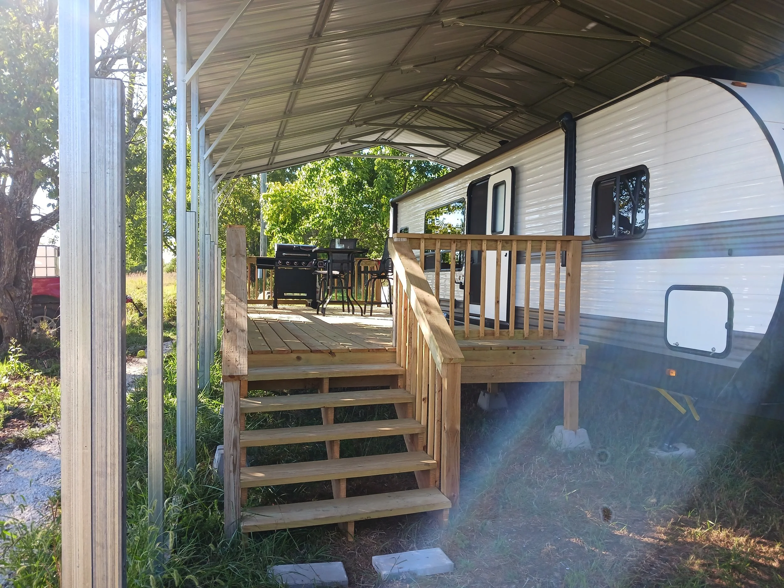 A white RV trailer with a wooden deck and stairs under a metal carport, with outdoor furniture and a grill on the deck, surrounded by greenery and trees.