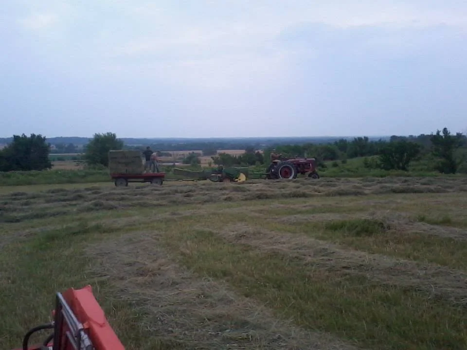 Farmland with hay being harvested, tractor, and farm workers on a cloudy day.