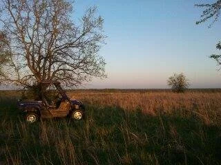 A small off-road vehicle parked in a grassy field under a tree during sunset or sunrise, with an open landscape and clear sky in the background.