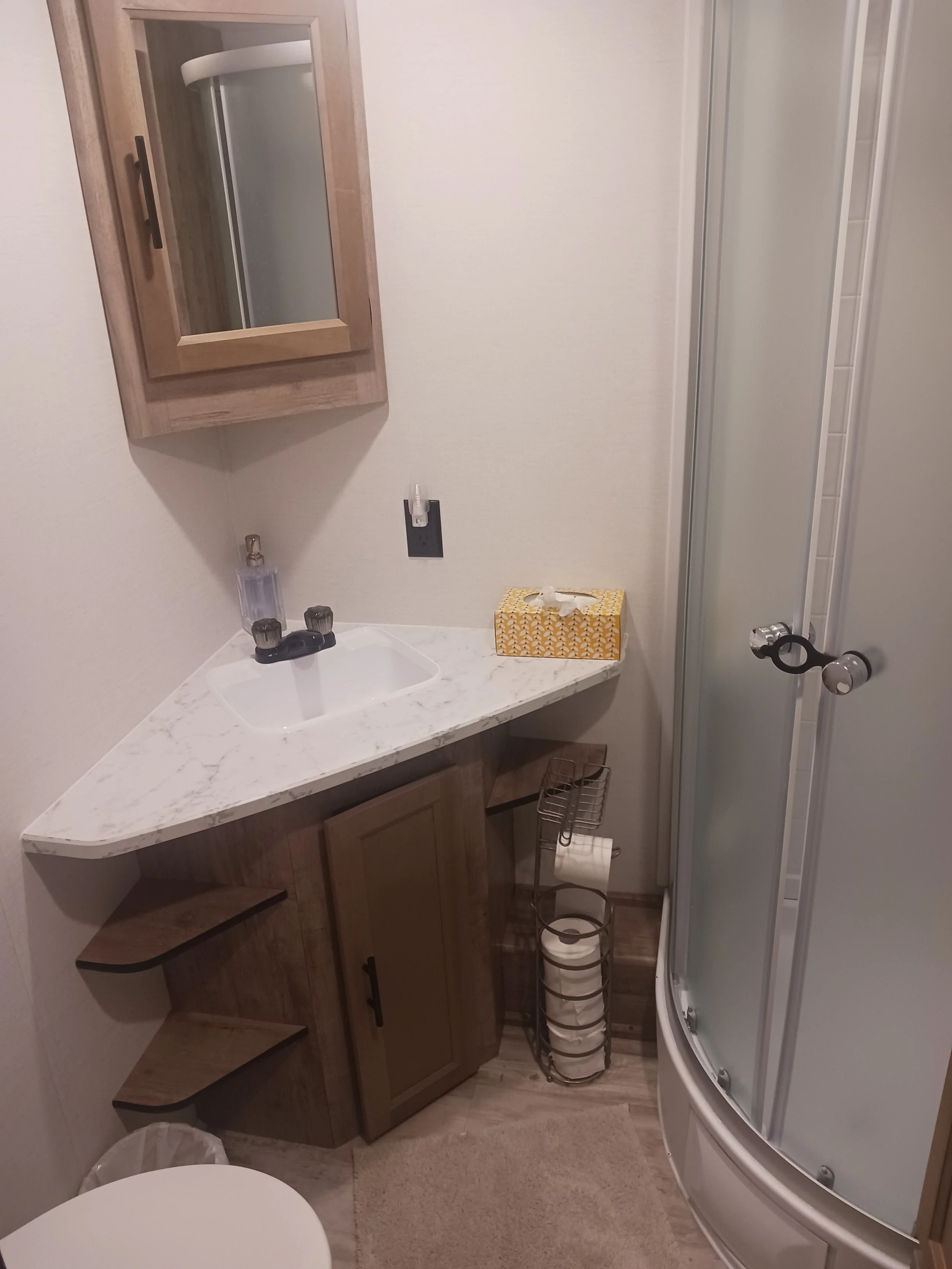Small bathroom with corner sink, wooden cabinet, mirror, tissue box, shower stall, toilet, and metal toilet paper holder with extra rolls.