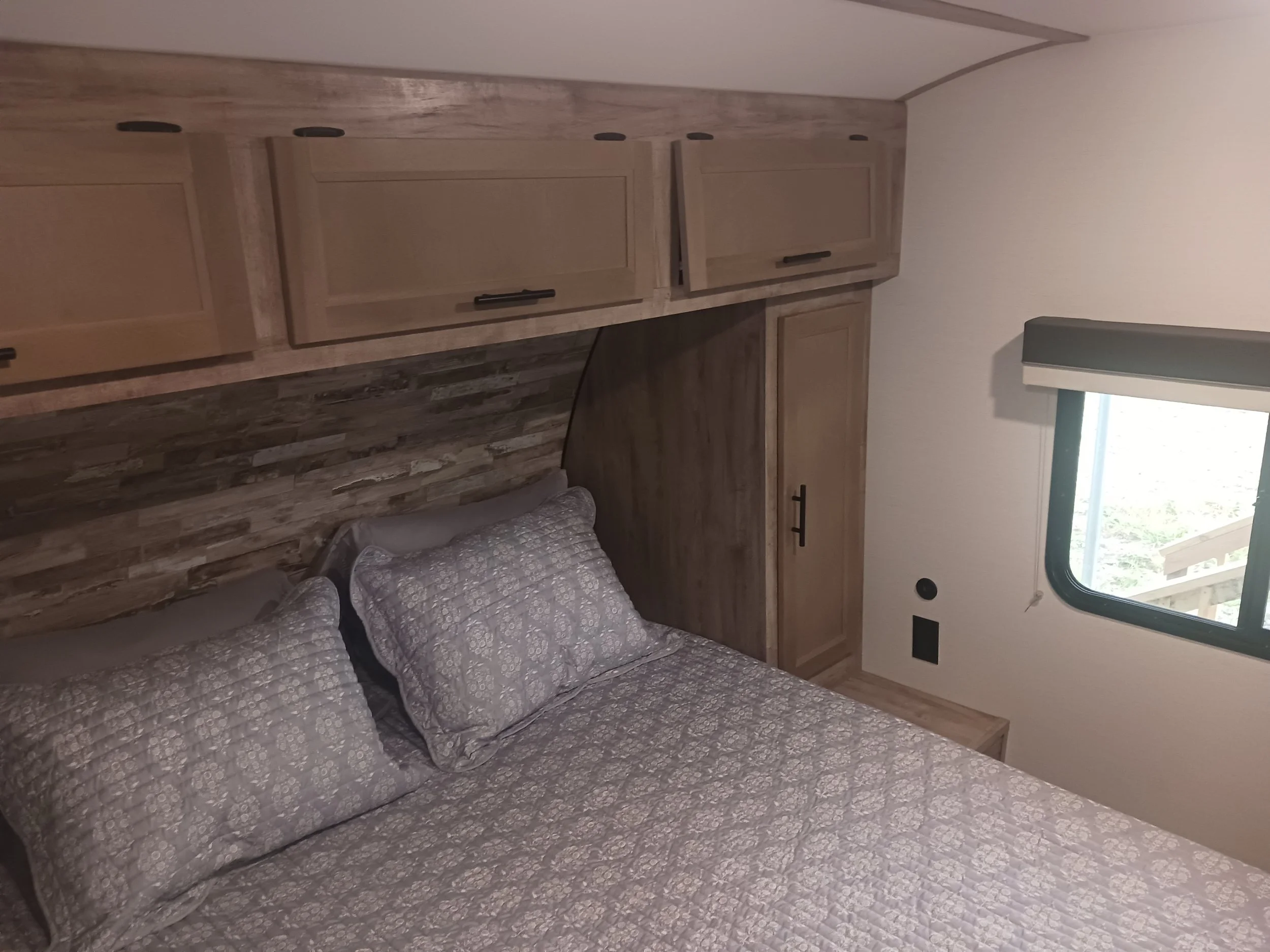 A bedroom with a bed, gray patterned bedding, and pillows, featuring a wood-paneled headboard and overhead cabinets, with a window on the right side.