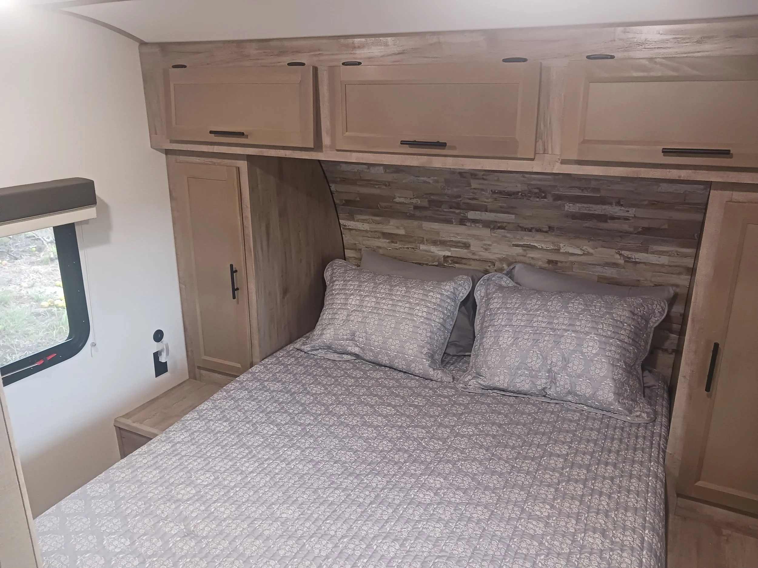 A bedroom with a bed covered in a patterned gray bedspread and matching pillows, a wood-paneled headboard with a stucco-like wall behind it, and built-in wooden cabinets above and beside the bed. There is a window with a black frame to the left.