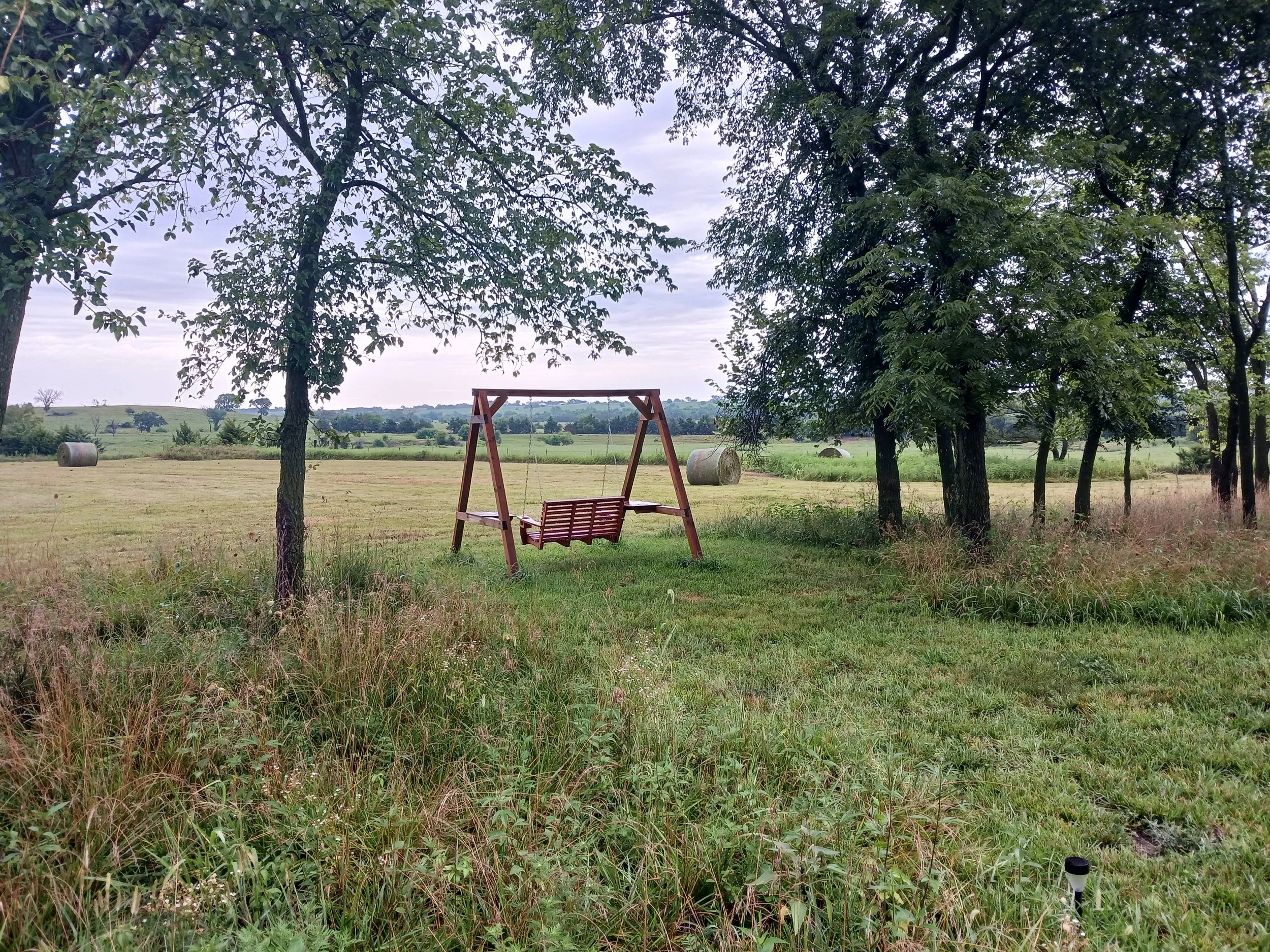A wooden swing bench hanging from a wooden frame on a grassy field surrounded by trees, with hay bales in the background and a cloudy sky overhead.