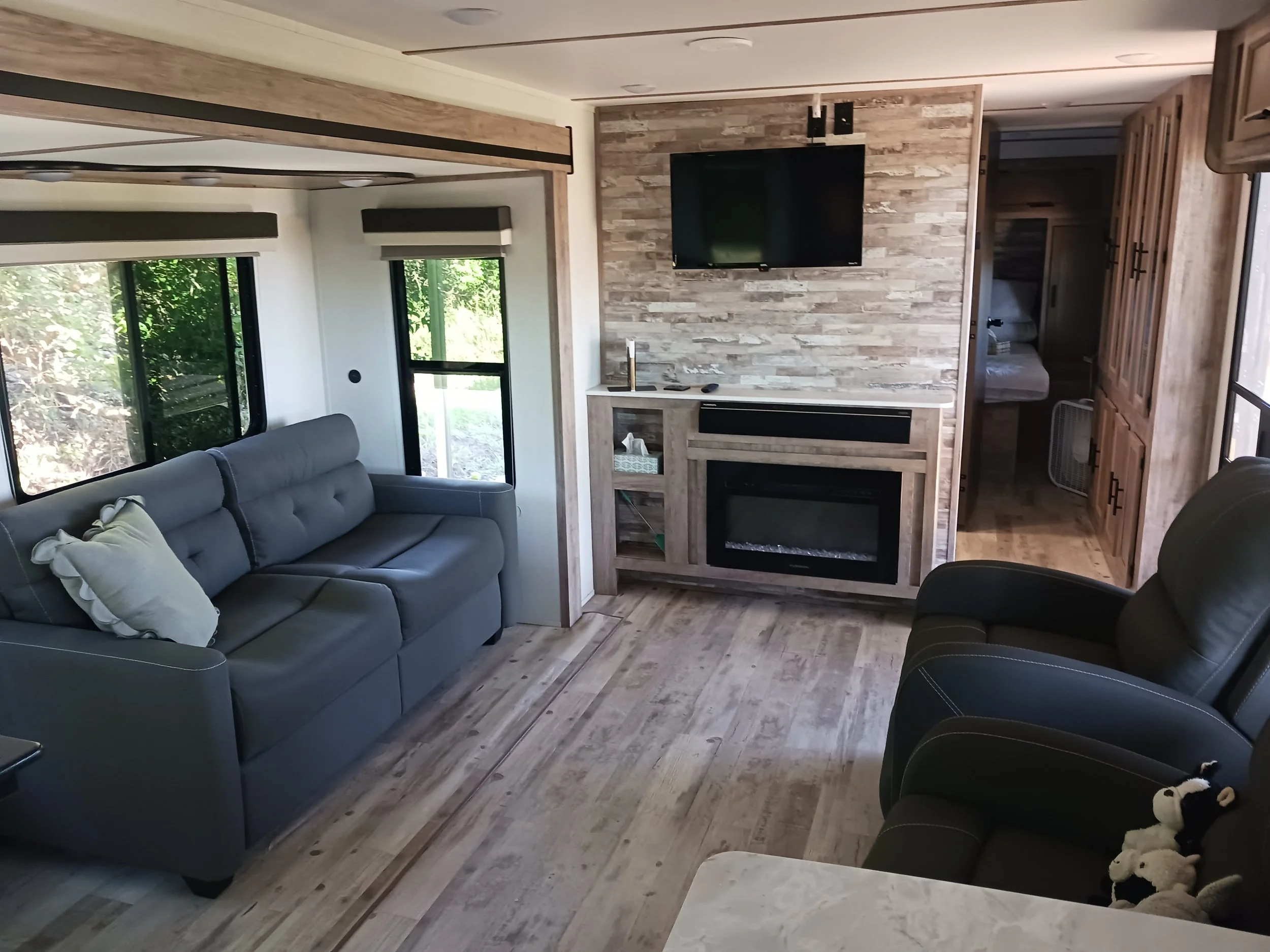 Living room with a gray sofa, a black recliner, and a fireplace with a mounted TV on a wooden accent wall. Large windows with views of greenery, and a doorway leading to a bedroom with a fan.