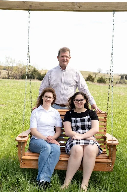 A man standing behind, smiling, with two women sitting on a wooden swing in a grassy field with trees and rolling hills in the background.
