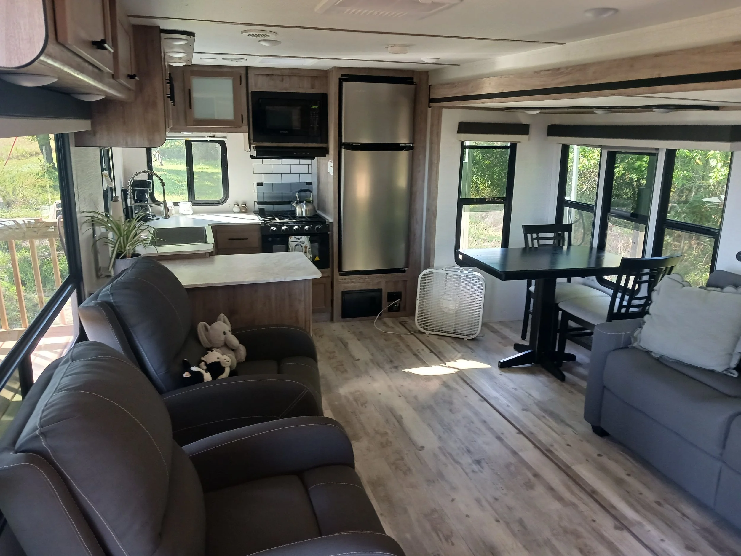 Interior of a cozy living space with a kitchen area, black armchairs with stuffed animals, a dining table with chairs, large windows with view of greenery, and a wood floor.