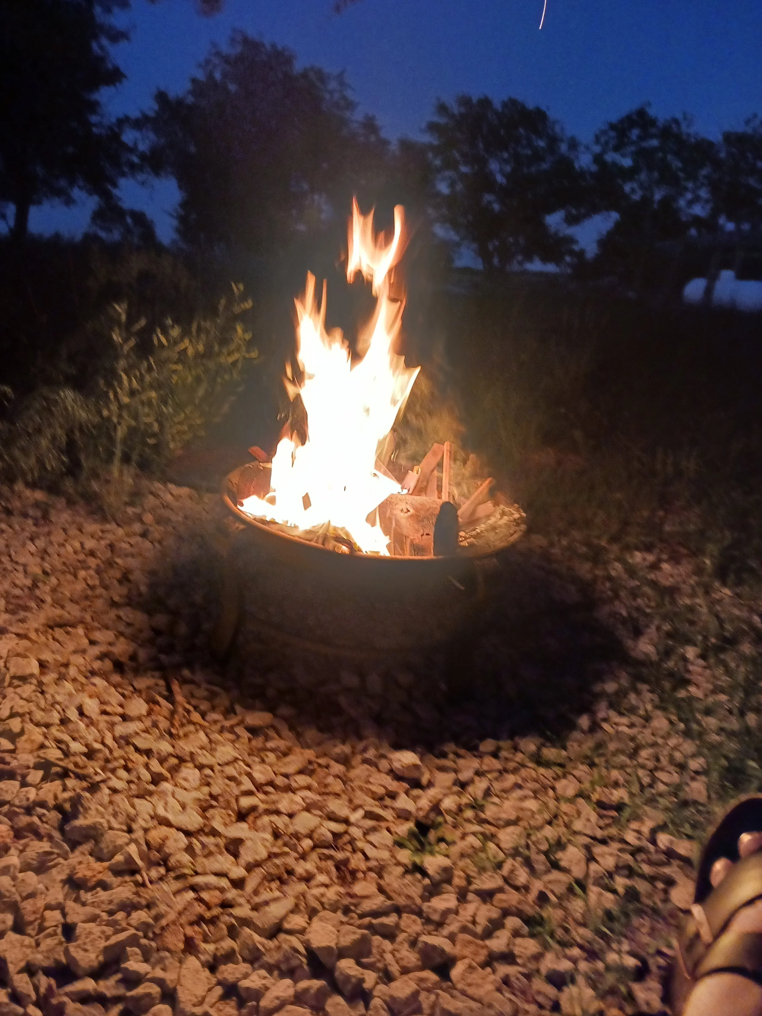 Campfire burning in a metal fire pit outdoors at night, surrounded by rocks and trees in the background.