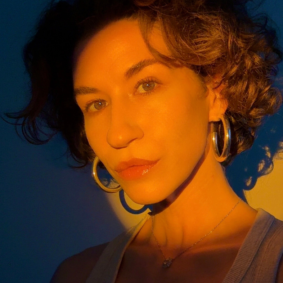 Close-up of a woman with short, curly hair, wearing hoop earrings and a necklace, illuminated by warm sunlight against a blurred background.