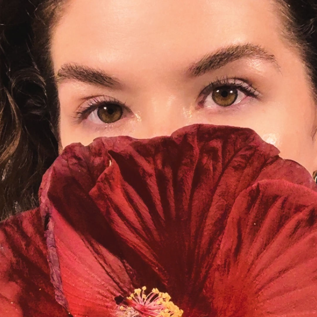 Close-up of a woman's face with brown eyes, partially covered by a large red flower she is holding up to her face.