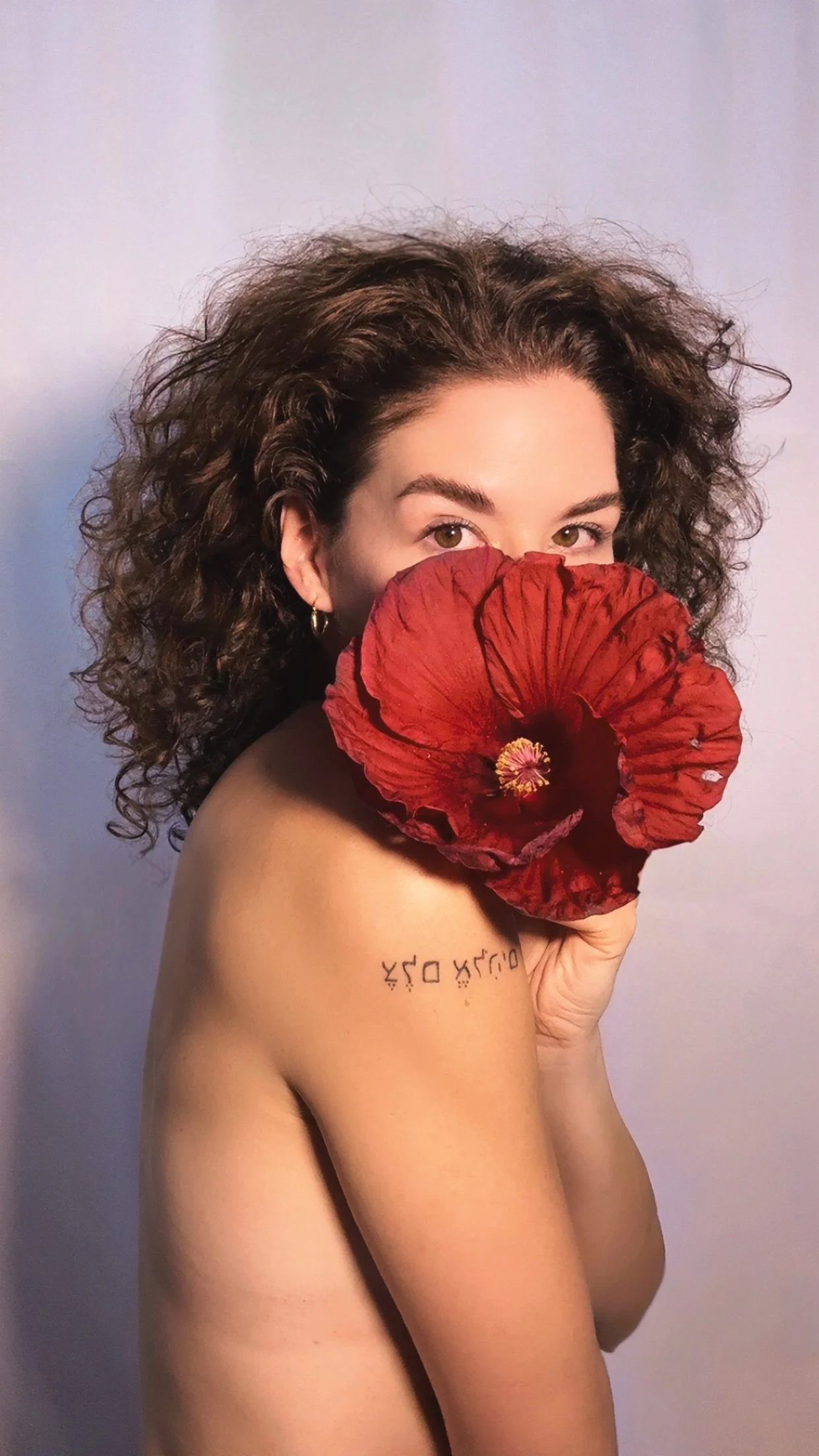 A woman with curly hair holding a red flower in front of her face, partially covering her mouth and nose, looking at the camera, with a tattoo on her shoulder.