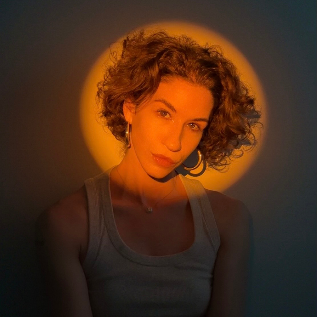 A woman with short curly hair wearing a tank top, earrings, and a necklace, illuminated by warm lighting with a circular glow behind her