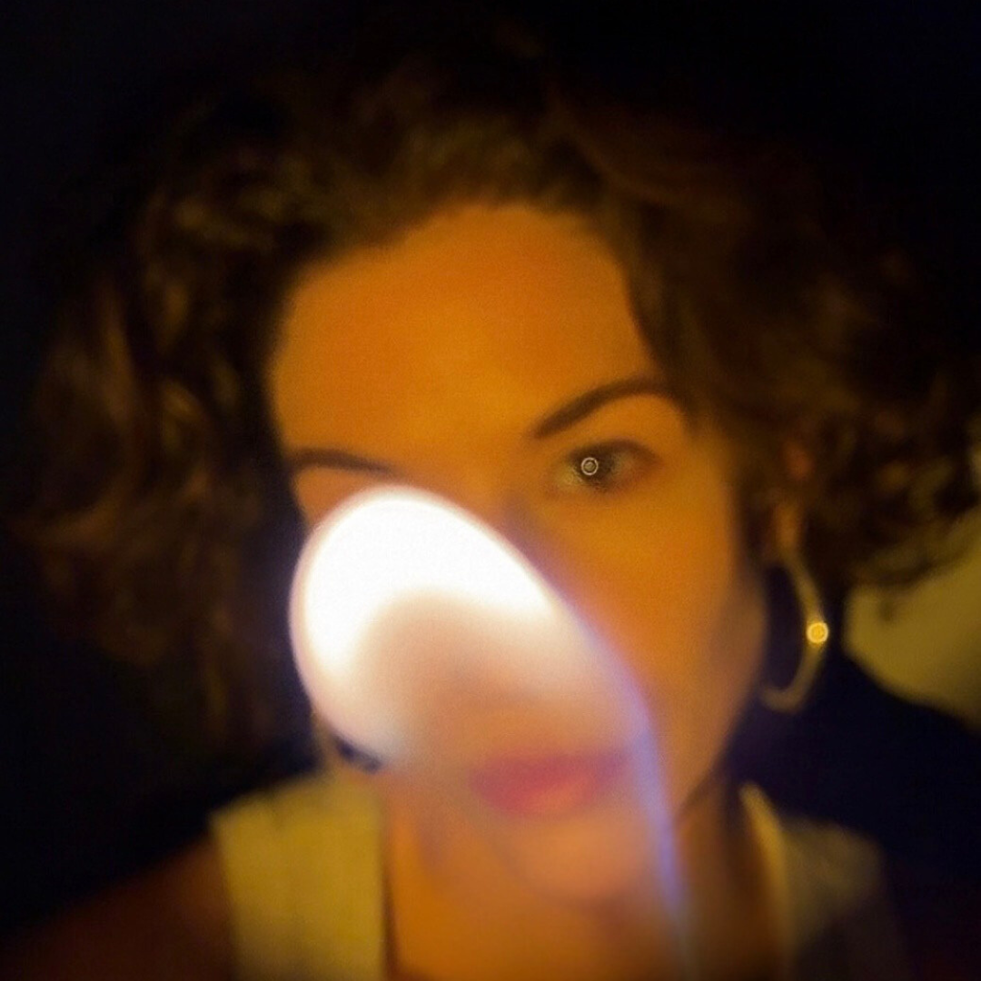 A woman with curly hair and hoop earrings holding a lit candle close to her face in a dark setting.