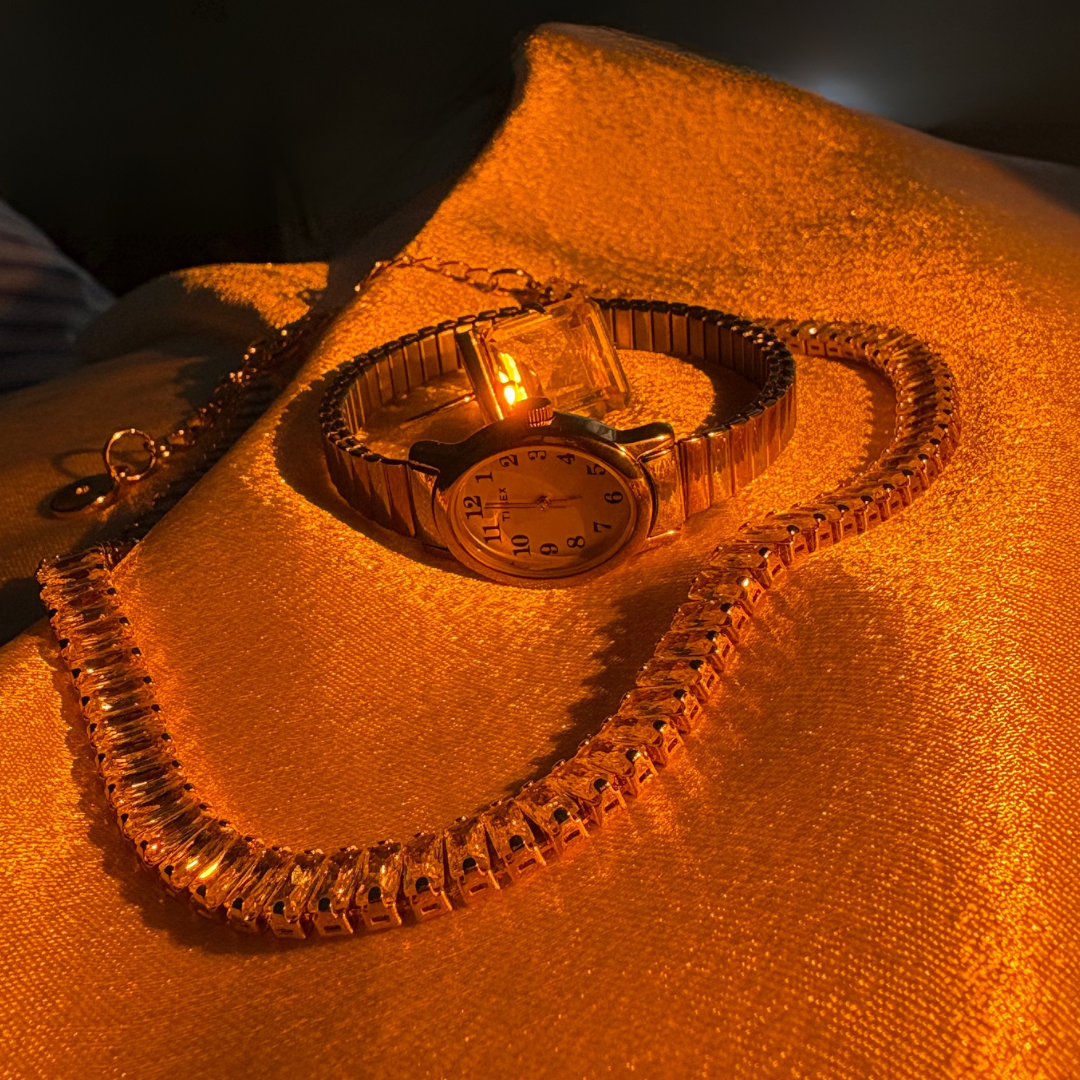 A gold wristwatch, a gold necklace, and a gold bracelet placed on a shimmery orange fabric.