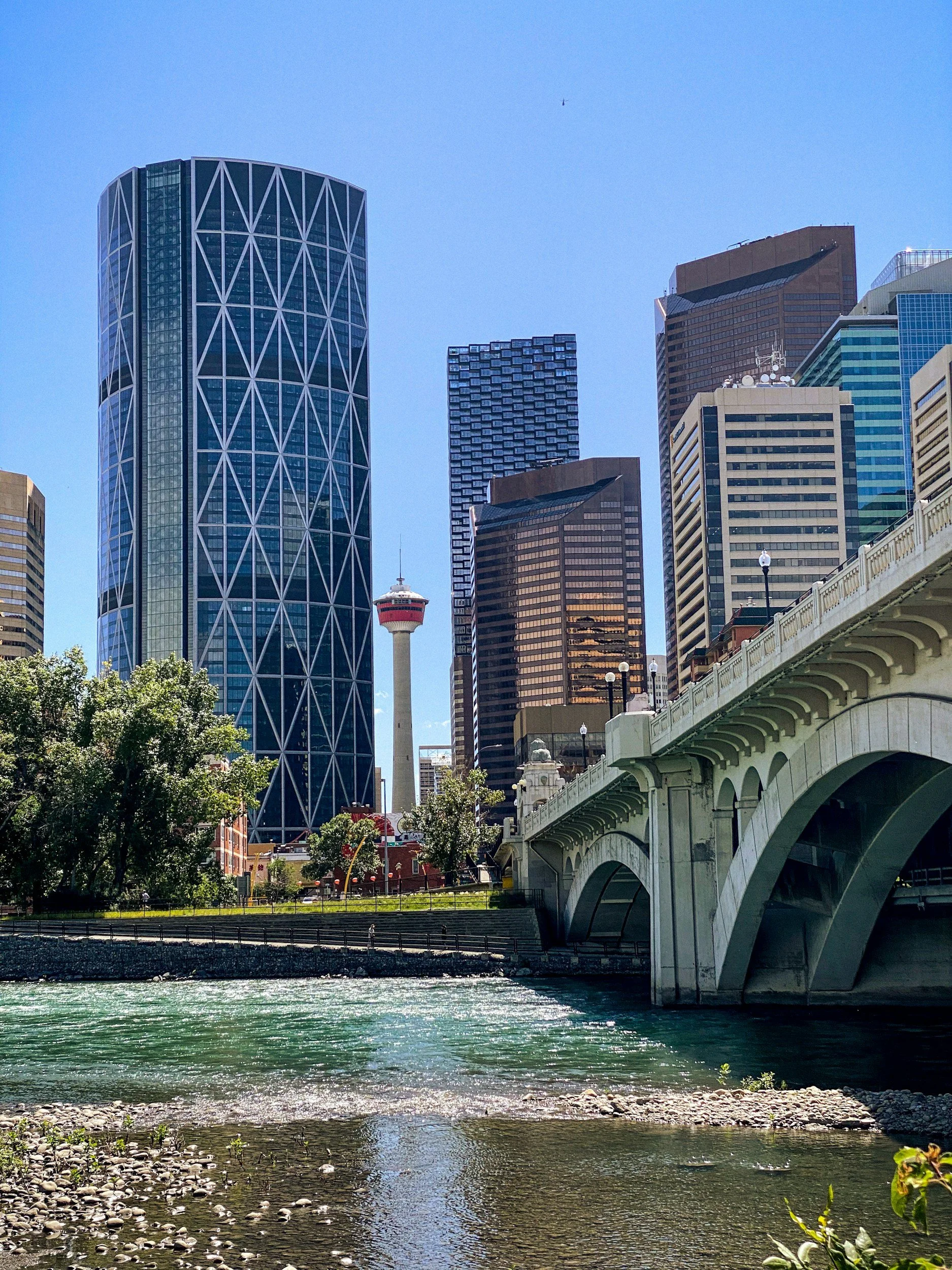 View of downtown Calgary with skyscrapers, the Calgary Tower, a river in the foreground, and a bridge crossing over the river.