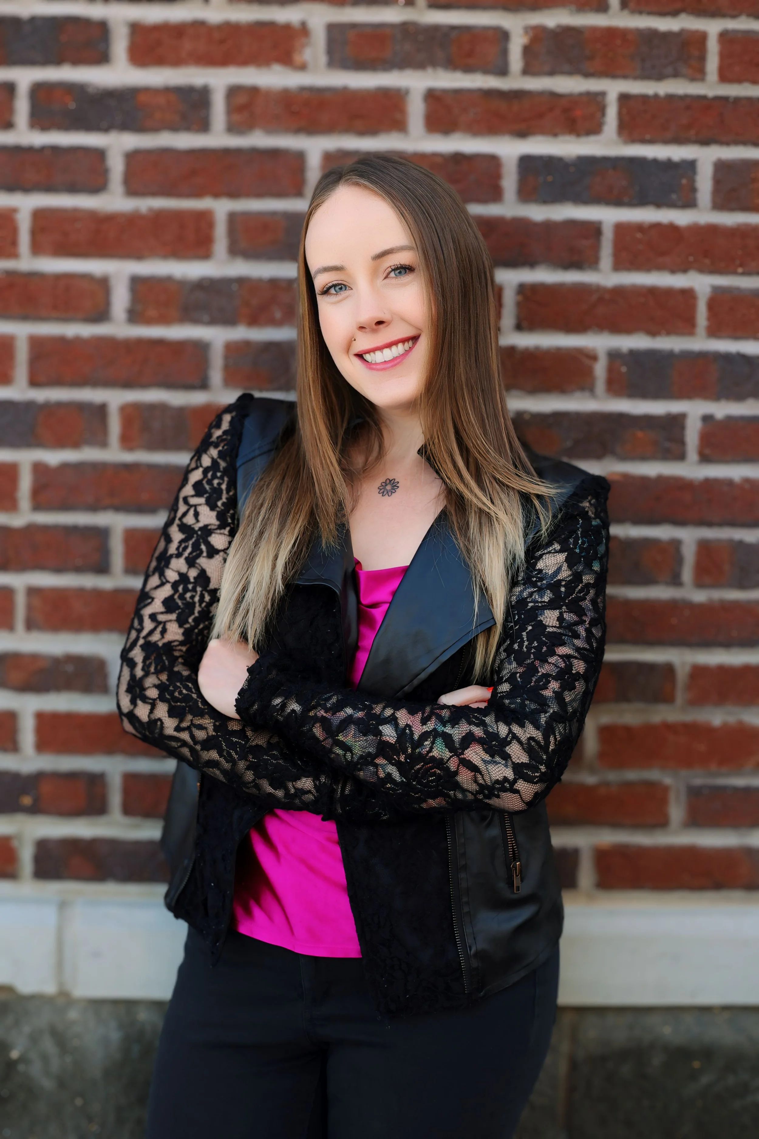 A young woman with long brown hair, wearing a black lace jacket, pink top, and black pants, stands with arms crossed in front of a red brick wall, smiling.