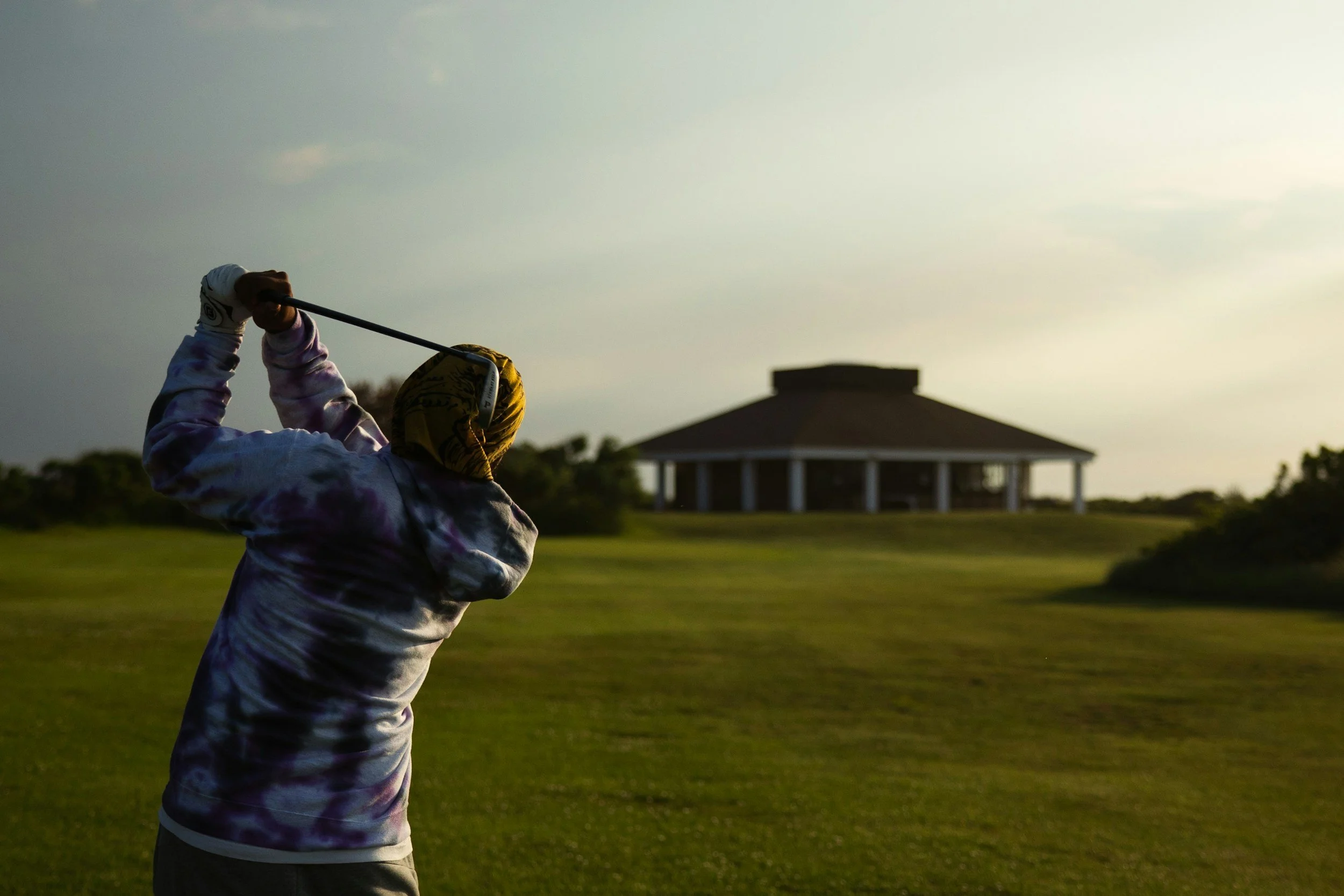 Person wearing a hoodie and yellow head covering swinging a golf club on a golf course with a pavilion in the background at sunset.
