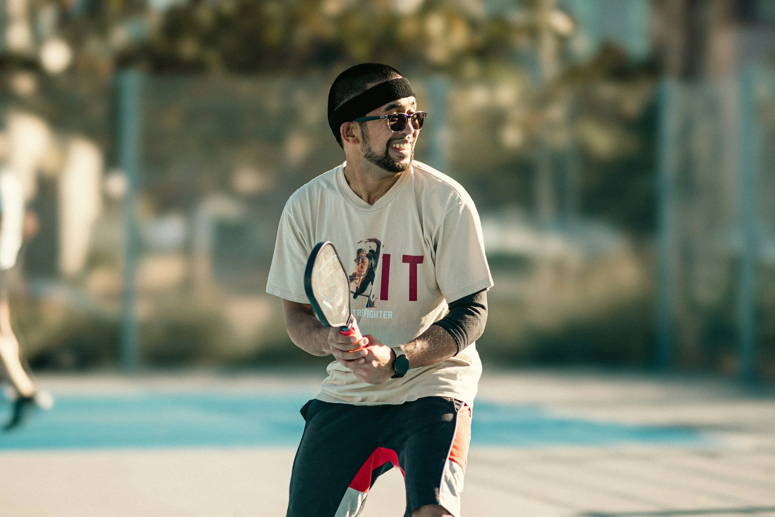 Person playing pickleball outdoors, wearing sunglasses, a black headband, a light-colored T-shirt, and black sports shorts.