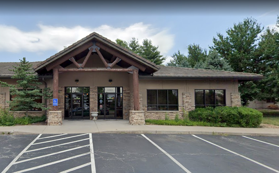 Exterior of a single-story office building with large windows, brick and wood facade, surrounded by trees and bushes, with parking spaces in front, including a handicap accessible spot.