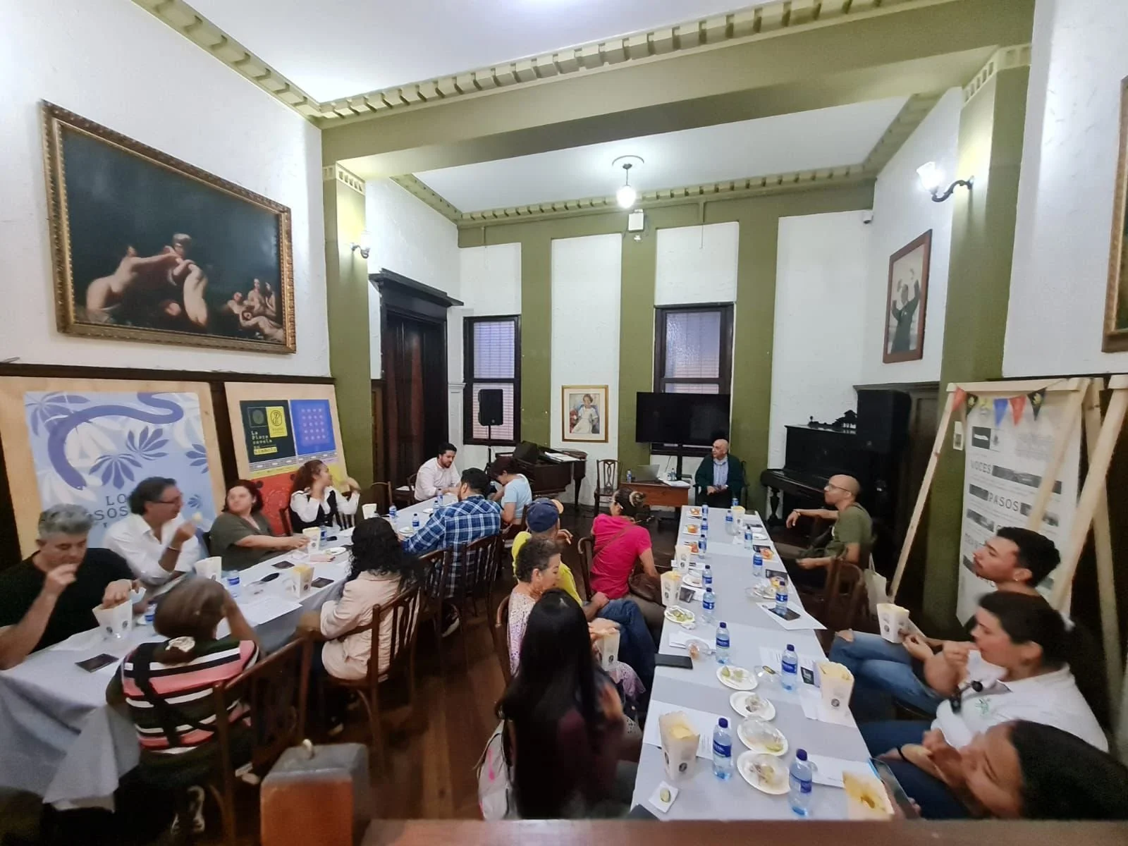 Personas en una cátedra o taller en una sala con paredes verdes, cuadros y ventanas, algunas oyendo, otras con botellas de agua y notas sobre la mesa.
