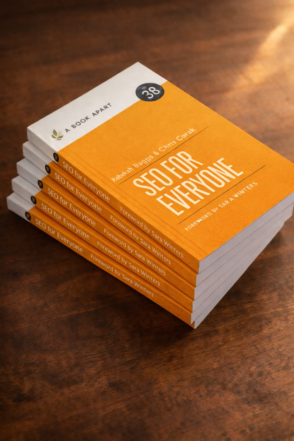 Stack of eight orange and white books titled 'SEO for Everyone' by Rebekah Baggs and Chris Corak, with a foreword by Sara Winters, on a wooden surface.