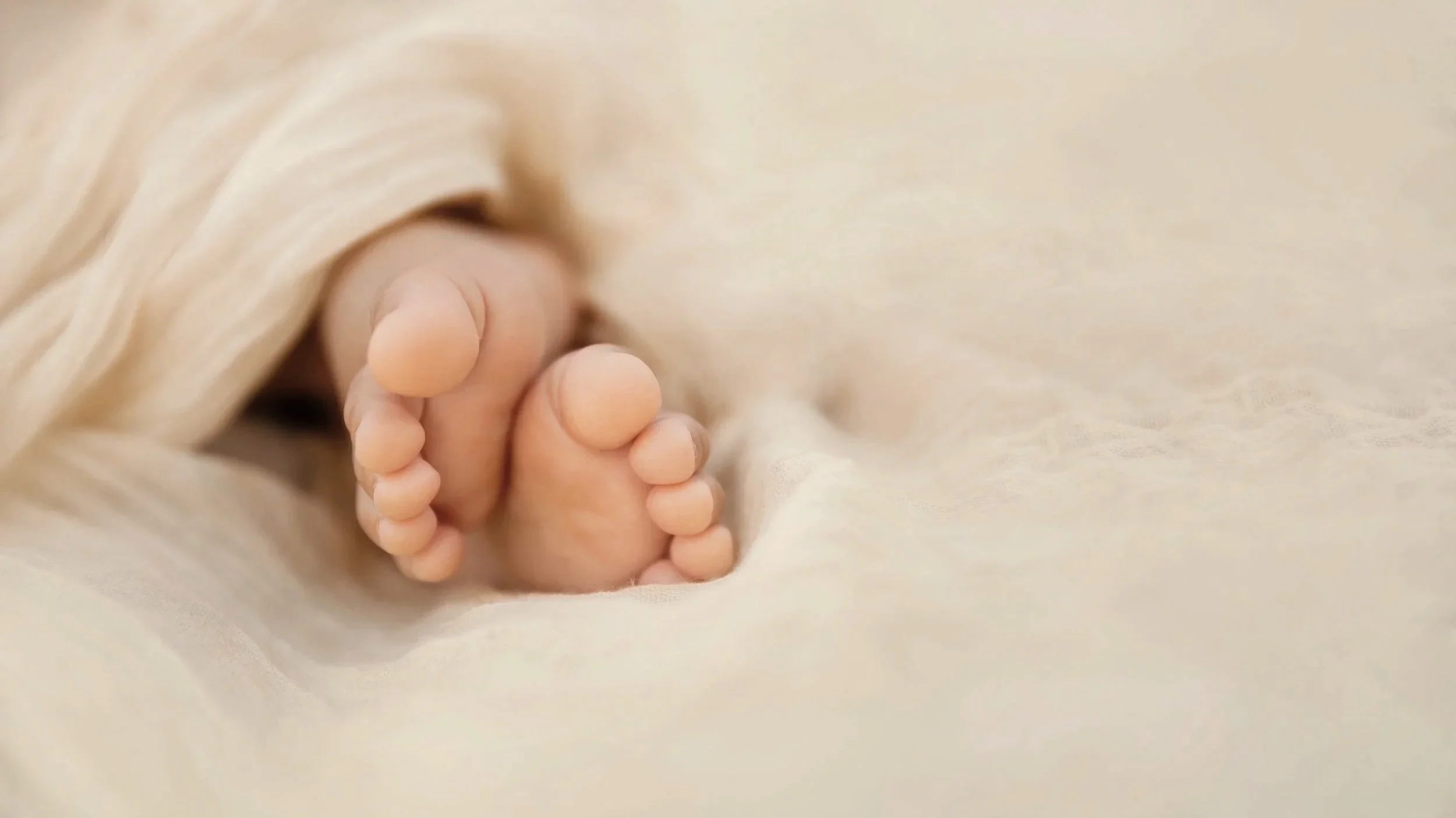 Petits pieds de bébé en gros plan, entourés de tissu beige, sur un fond clair et doux.