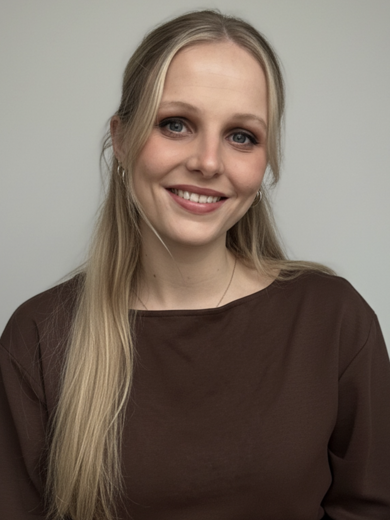 Jeune femme avec longs cheveux blonds, portant un haut marron, souriant devant un fond neutre.