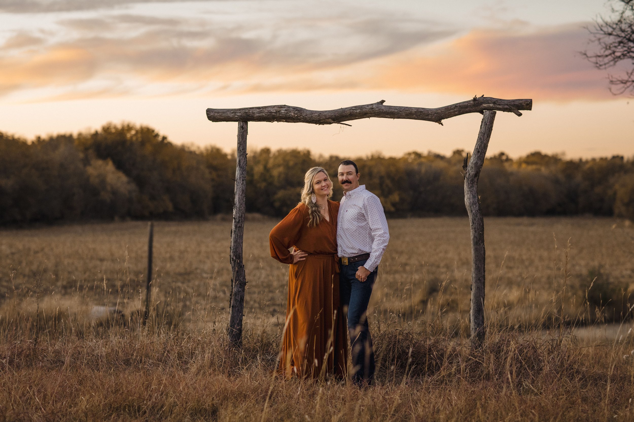 A couple standing together under a rustic wooden arch in an open field during sunset, with trees in the background.
